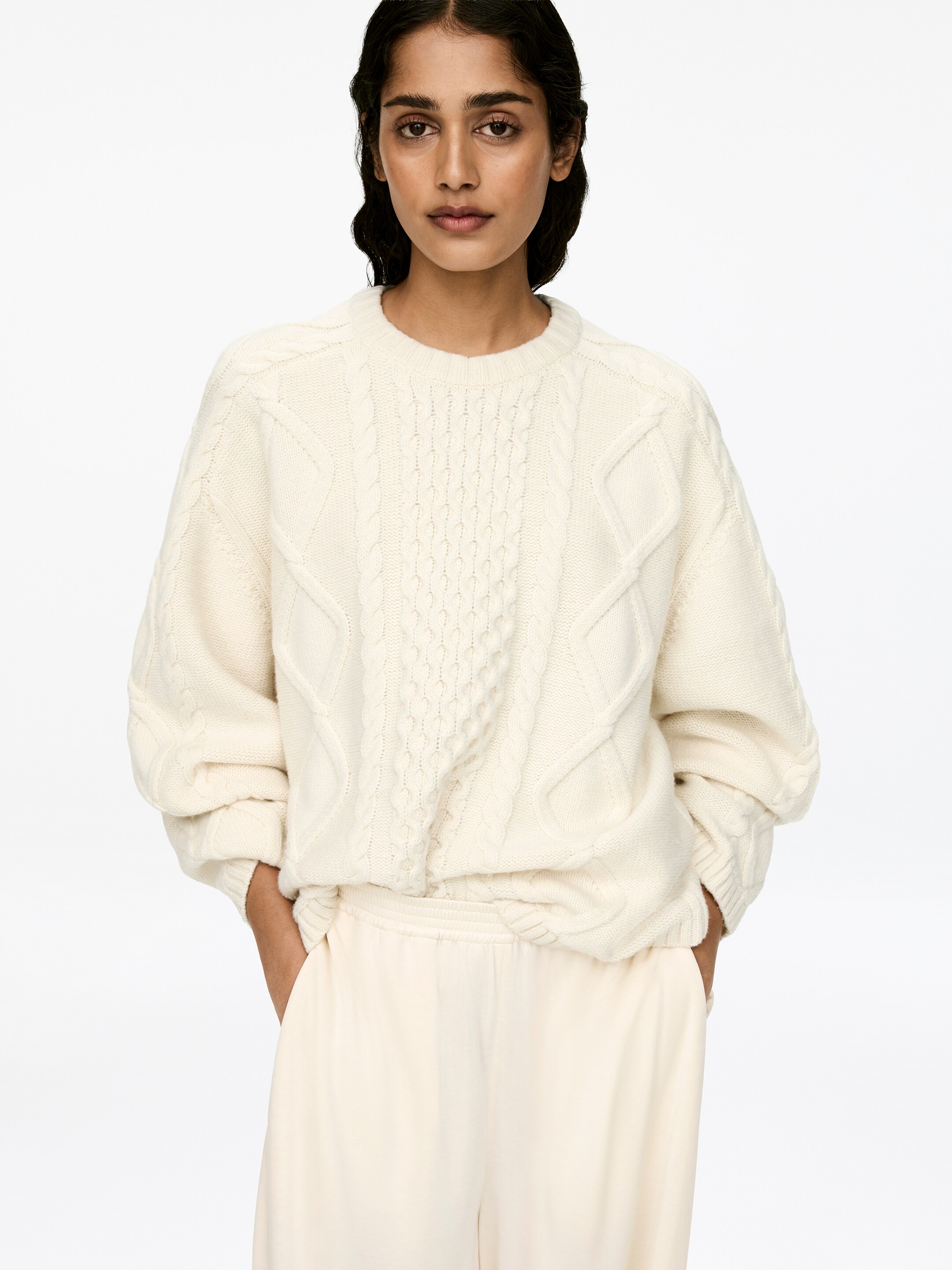 View larger image: A cream cable knit jumper with a crew-neck, long sleeves, and ribbed trims is being worn by the model. The model's hands are resting in pockets, illustrating the jumper's relaxed fit.