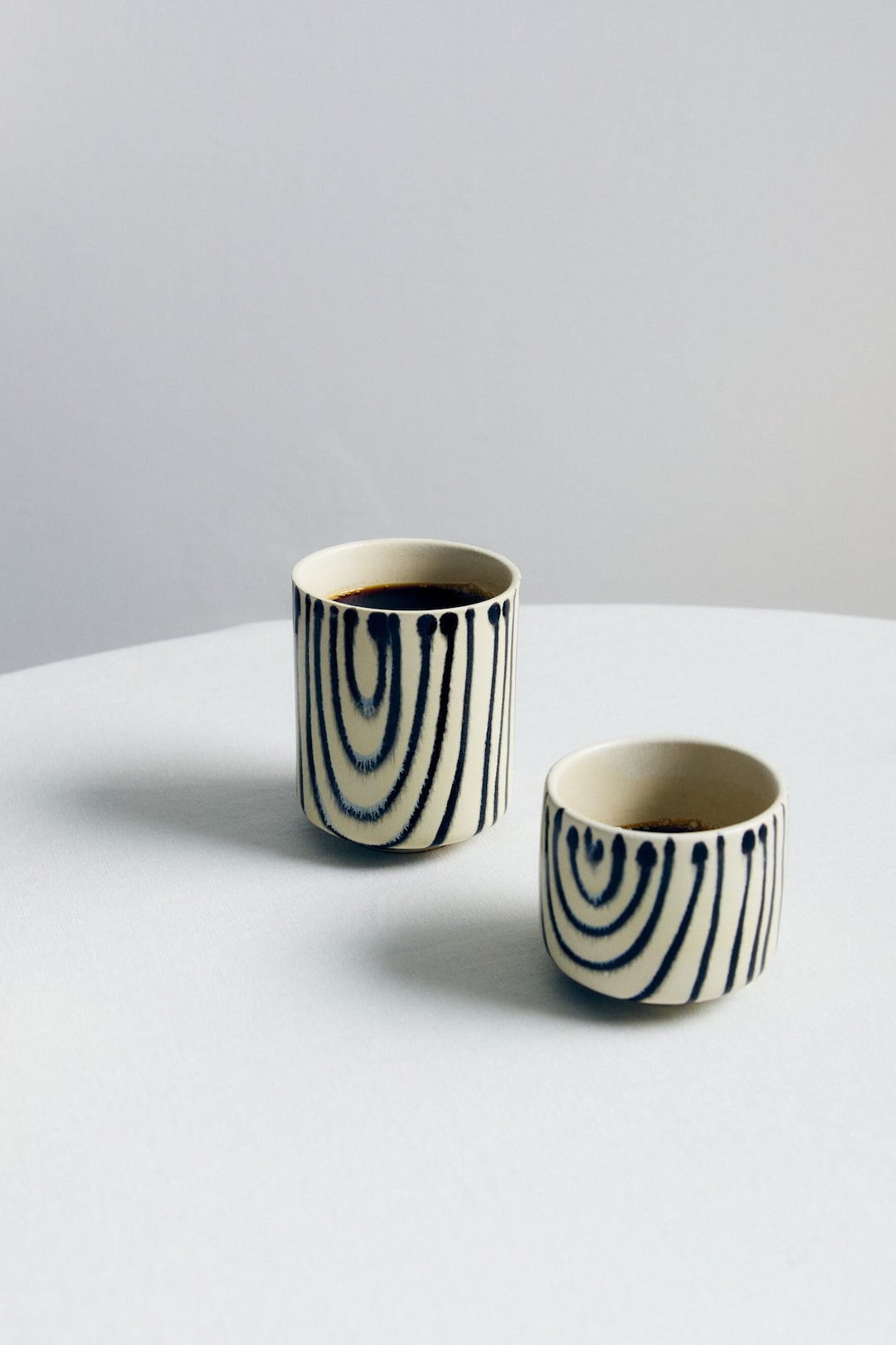 Ver imagen más grande: Tres tazas de cerámica presentan un color base claro con patrones oscuros, abstractos, de líneas verticales y curvas. Cada taza cilíndrica contiene un líquido oscuro, mostradas desde un ángulo frontal ligeramente elevado.