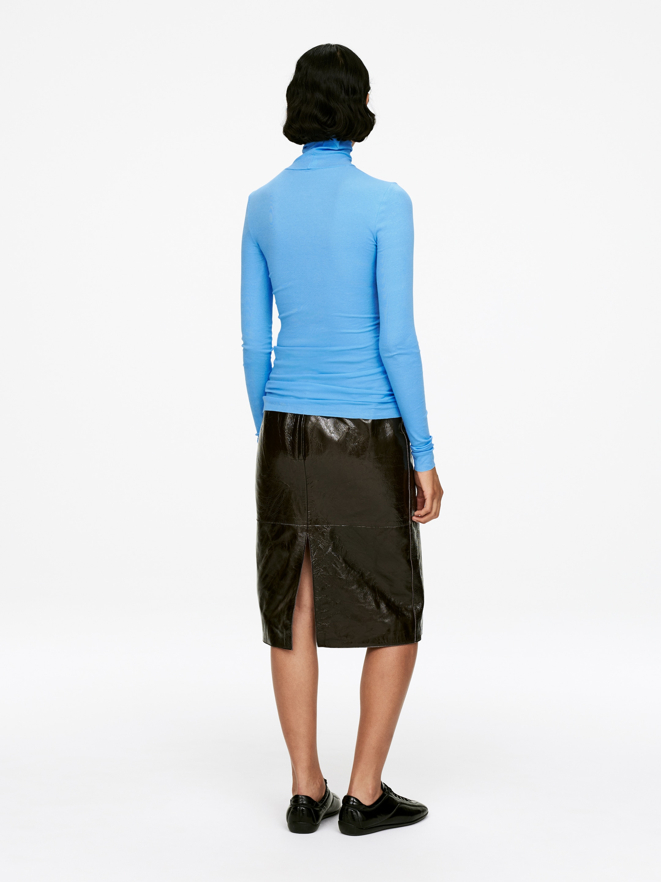 Agrandir l'image: Un haut à col roulé bleu clair et ajusté est porté de dos, avec des manches longues et un col haut. Il est associé à une jupe foncée, brillante, longueur genou, avec une fente à l'arrière et un panneau visible.