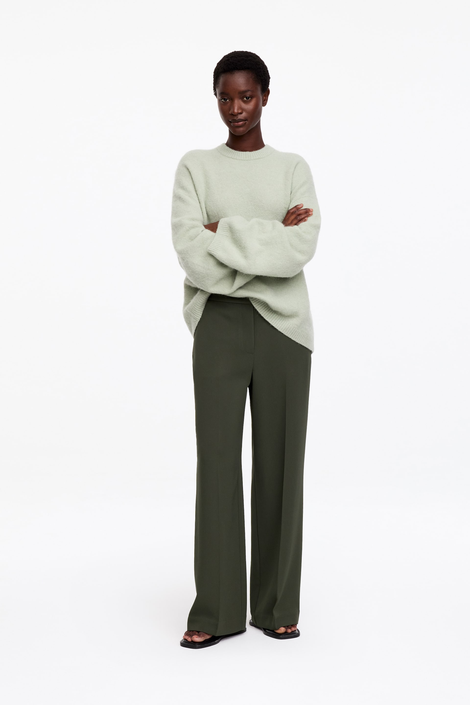 Grotere afbeelding bekijken: De model draagt een donkergroene, rechte broek met geperste vouwen vooraan. De broek is gecombineerd met een lichtgroene gebreide trui met lange mouwen en ronde hals.