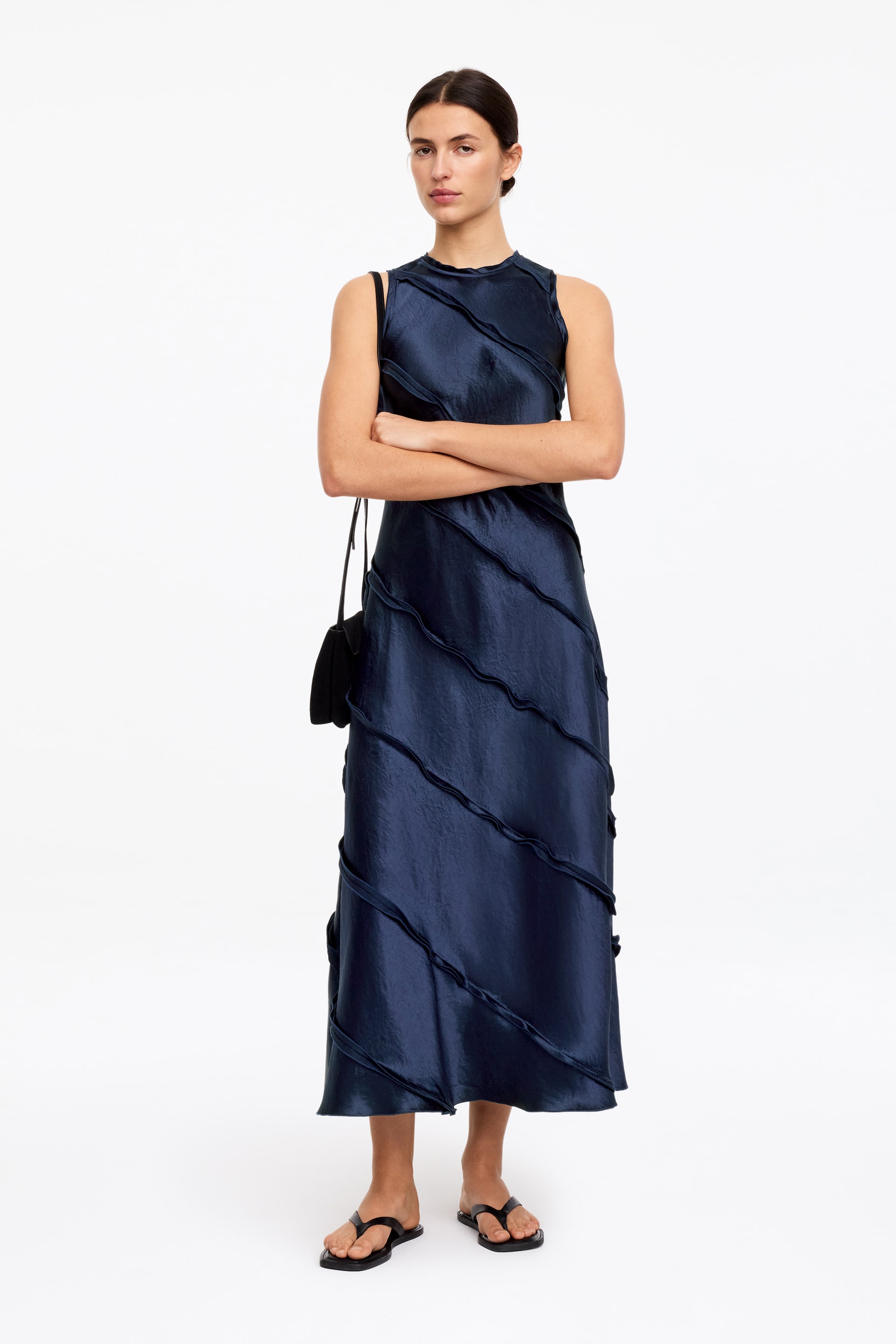 View larger image: A dark blue sleeveless midi dress features diagonally stitched fabric strips across its front, creating a wavy, textured pattern. The dress presents a shimmery appearance and is worn with a black shoulder bag resting on its side.