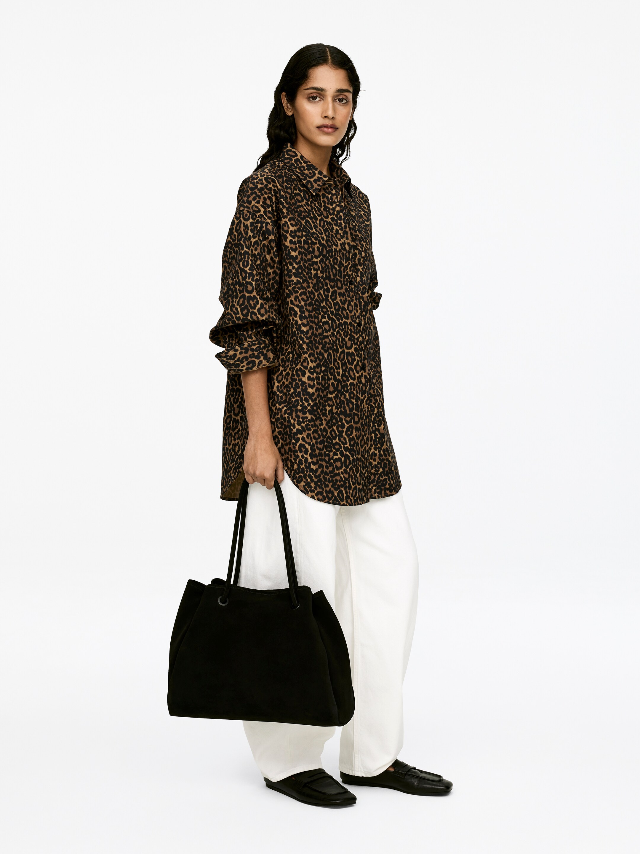 Größeres Bild ansehen: Ein übergroßes Hemd mit Leopardenmuster, das über einen Kragen und Fledermausärmel verfügt, wird von dem Model getragen. Das Model trägt außerdem weiße Hosen mit weitem Bein und trägt eine schwarze Tragetasche.