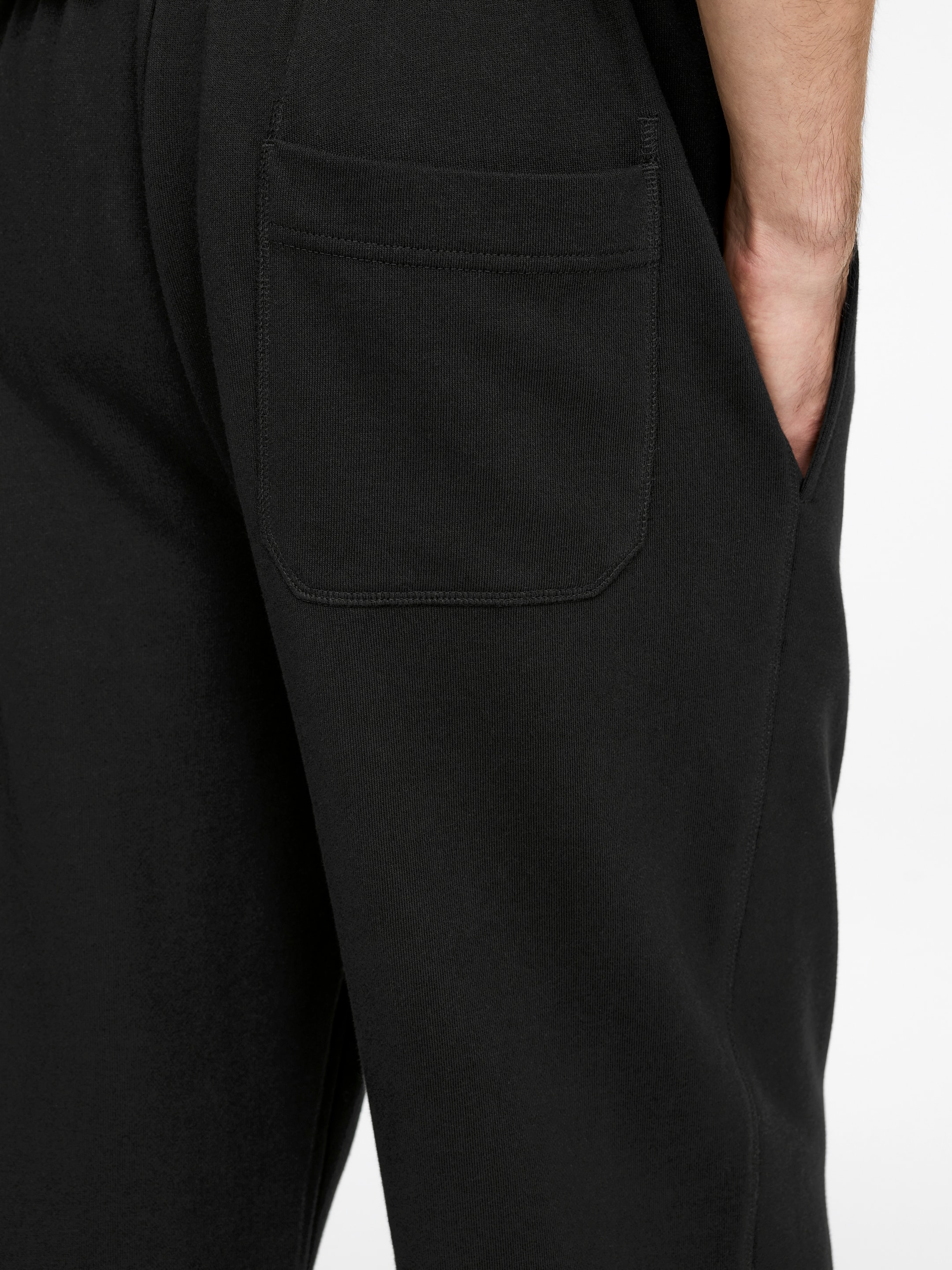 Größeres Bild ansehen: Schwarze Jogginghosen werden aus einem leicht schrägen, rückseitigen Winkel gezeigt, mit einer rechteckigen Patte am Gesäßtasche auf dem rechten Bein und einer Hand, die in einer Seitentasche steckt.