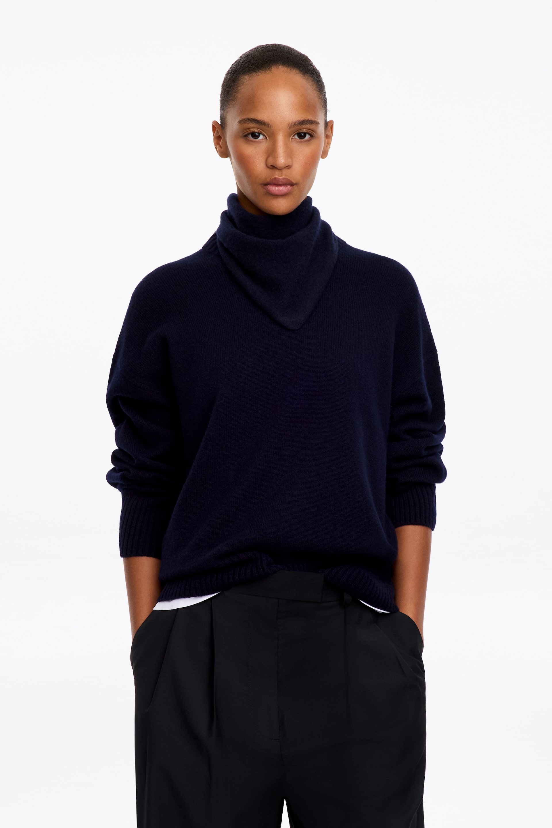 Visualizza immagine più grande: Un maglione in maglia blu navy con un alto collo a imbuto morbido e drappeggiato indossato dalla modella. Il maglione presenta maniche lunghe con polsini a costine ed è indossato sopra uno strato inferiore bianco, abbinato a pantaloni neri.