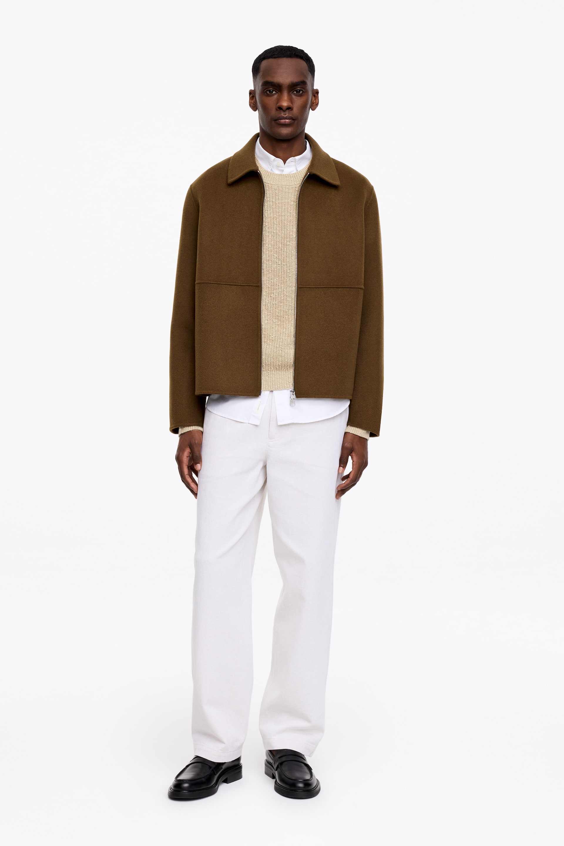 View larger image: A brown, short, boxy jacket is displayed from the front, featuring a spread collar, a full-zip fastening with a visible zip pull at the hem, and horizontal seam detailing across the chest. It is styled layered over a light beige crew-neck jumper and a white collared shirt.