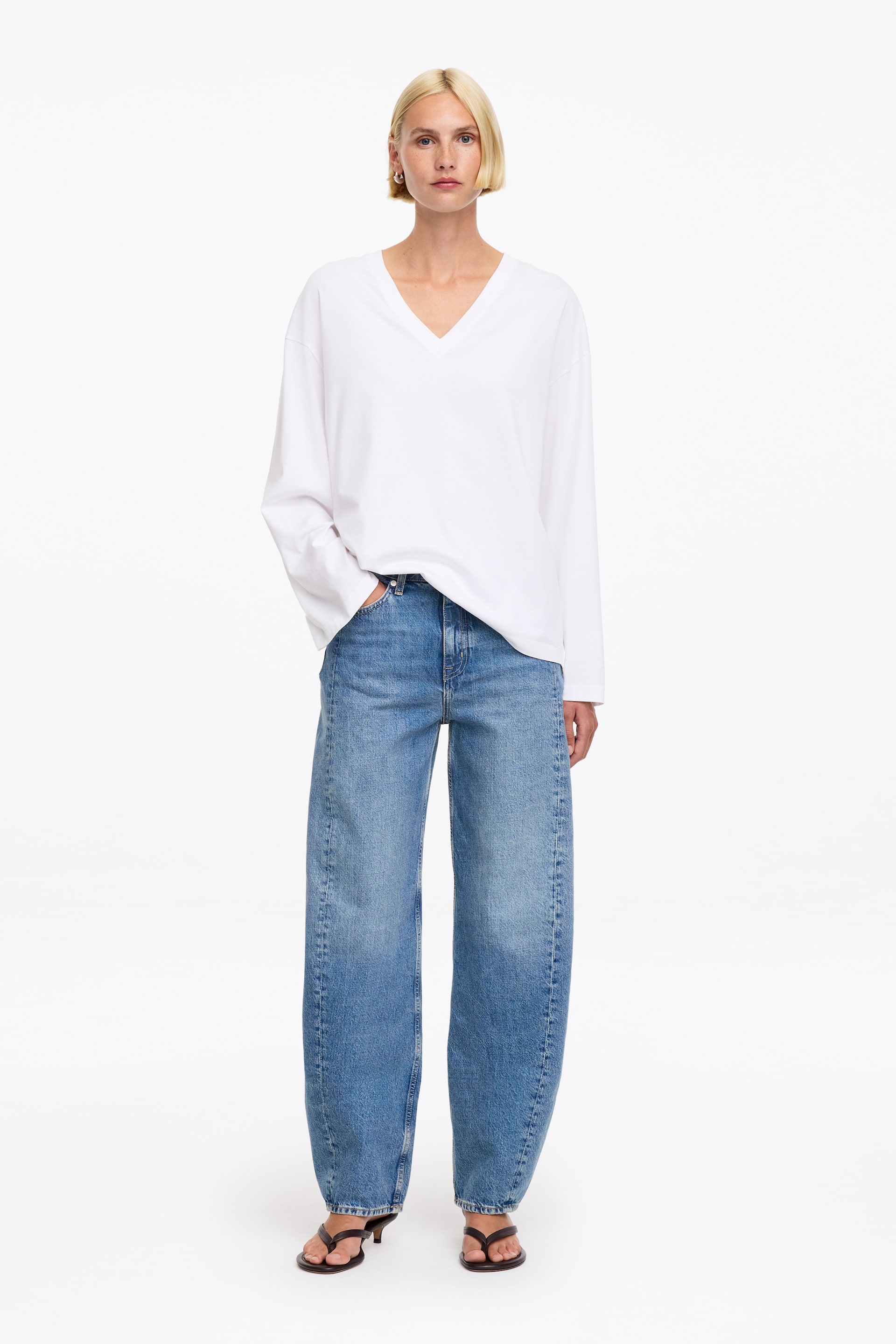 Grösseres Bild ansehen: Ein weißes, übergroßes V-Ausschnitt-Langarmshirt wird zusammen mit blauen, hoch taillierten, weit geschnittenen Jeans präsentiert, die sichtbare Frontnähte und eine lockere Passform aufweisen, alles in Frontalansicht. Dunkelbraune Zehensandalen mit Absatz werden zu diesem Outfit getragen.
