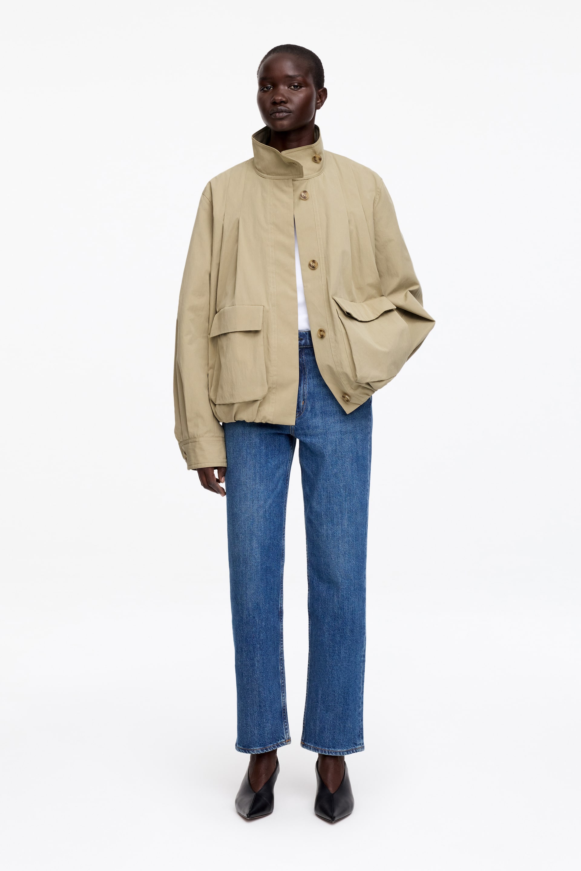 Grotere afbeelding bekijken: Een beige blousonjack wordt gedragen door een model en heeft een opstaande kraag met zichtbare knopen, een knoopsluiting aan de voorkant, verticale plooien aan de voorkant, grote opgestikte zakken en een geplooide bubbelszoom. De mouwen van het jack hebben geplooide, geknoopte manchetten, en het model draagt het over een witte top en blauwe jeans, met één hand rustend in een voorzak.