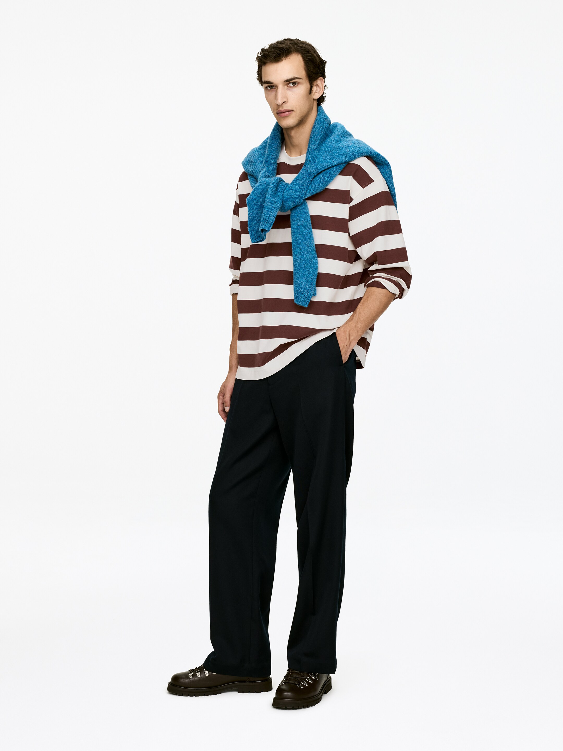 Agrandir l'image: Une personne est debout, légèrement inclinée vers la droite, portant une chemise à manches longues à rayures crème et marron ample, avec un pull texturé bleu drapé sur ses épaules. Elle porte également un pantalon large et sombre et des chaussures à lacets marron foncé avec des œillets argentés et des semelles épaisses.