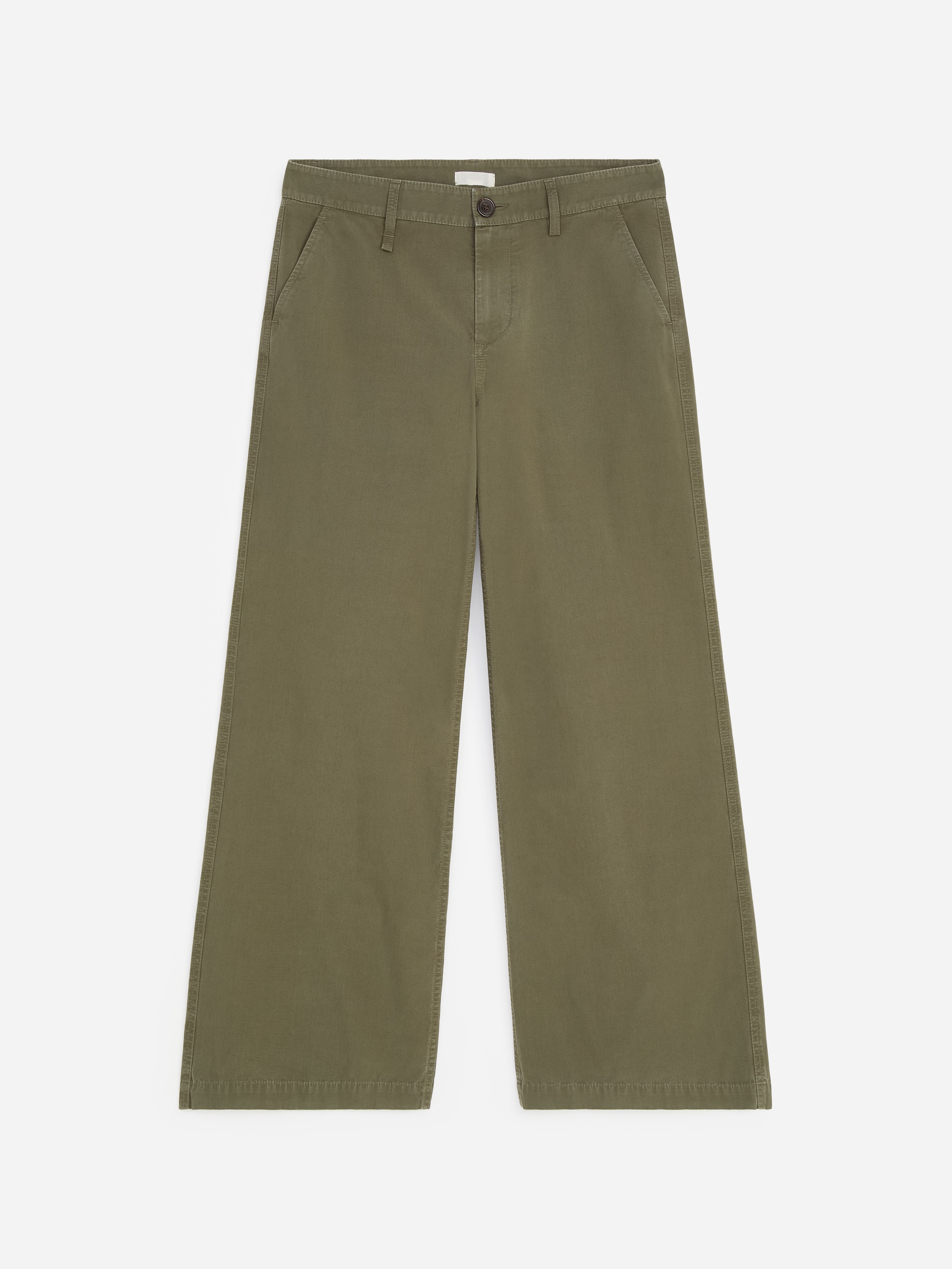La modella indossa pantaloni chino a gamba larga verde oliva, con le mani inserite nelle tasche laterali oblique. I pantaloni a vita bassa sono chiusi da una sottile cintura nera e abbinati a una camicia bianca a maniche lunghe infilata dentro.