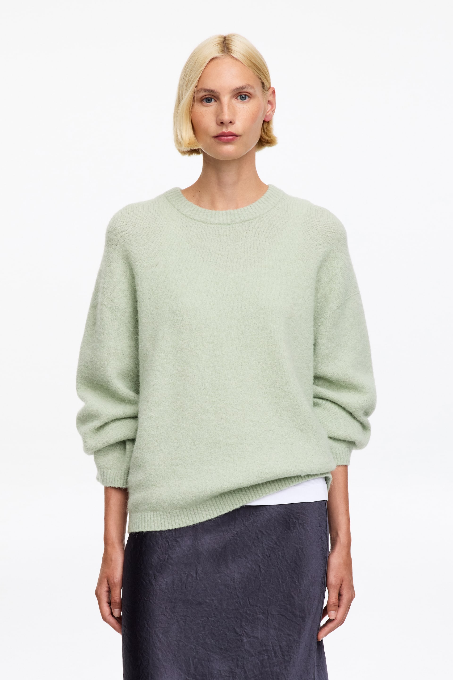 View larger image: A light green knitted jumper, featuring a fuzzy finish, an oversized fit, and ribbed trims, is being worn by the model. A white undershirt is visible beneath the jumper, which is being styled with a dark skirt.