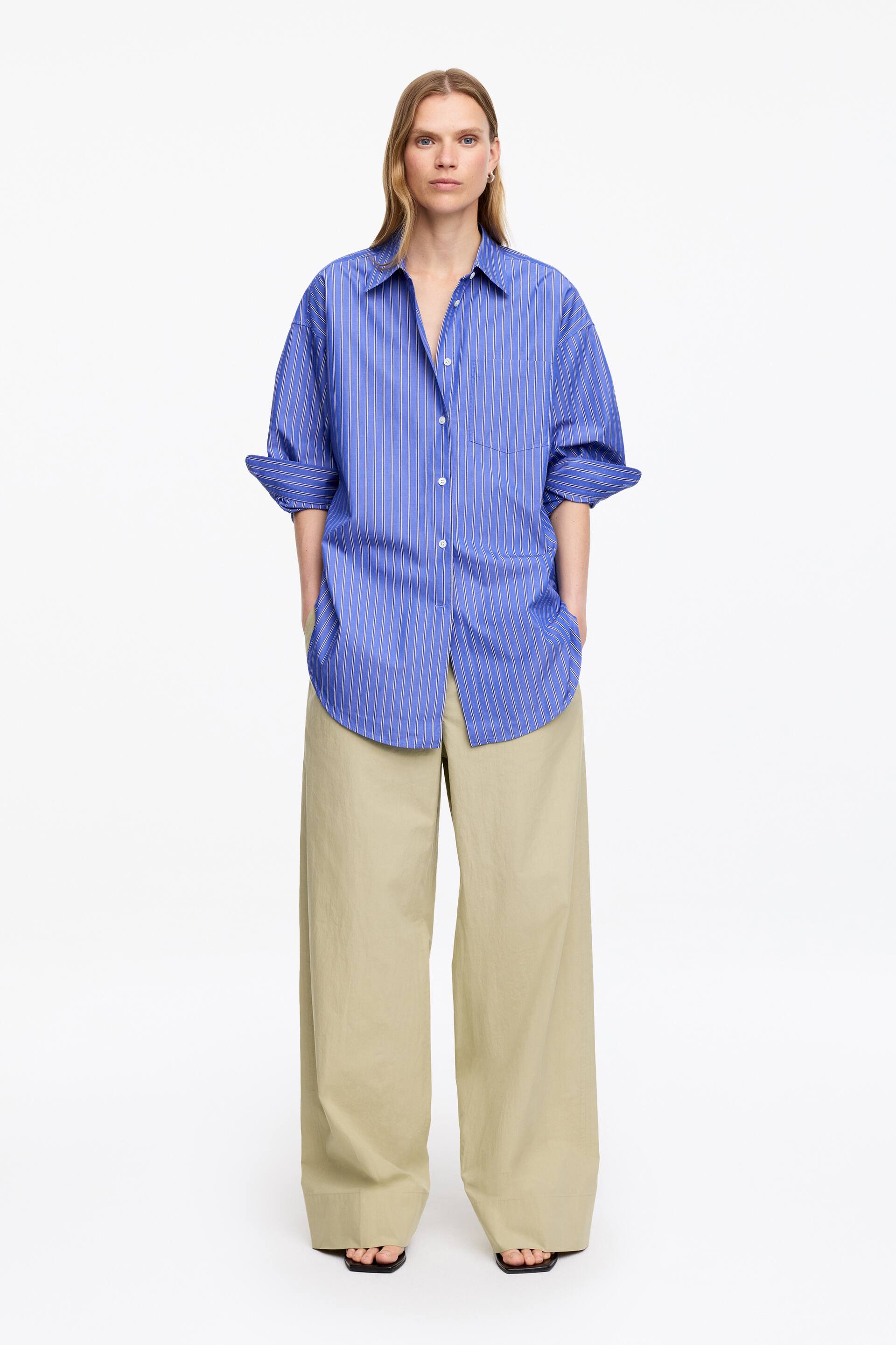 Ver imagen más grande: La camisa de rayas azules presenta finas rayas verticales blancas y un corte holgado. Está diseñada con un cuello en punta, una parte delantera con botones, un único bolsillo de parche en el pecho izquierdo y sus mangas largas están arremangadas.