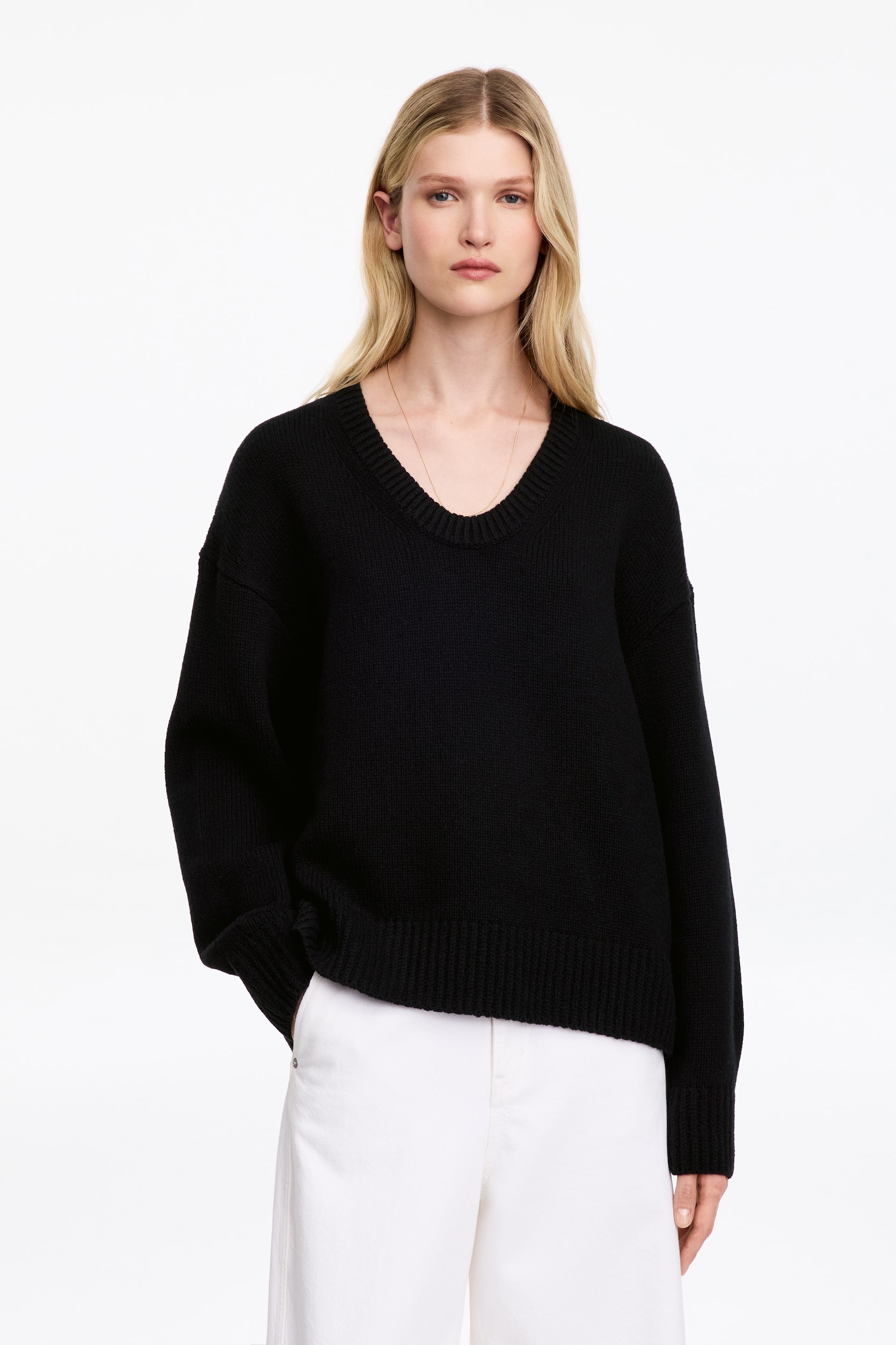 Visualizza l'immagine ingrandita: Un maglione nero in maglia con profondo scollo a U a coste, spalle scese e bordi a coste è indossato dalla modella. Il maglione dalla vestibilità morbida è abbinato a pantaloni bianchi, mentre la modella tiene una mano in tasca.