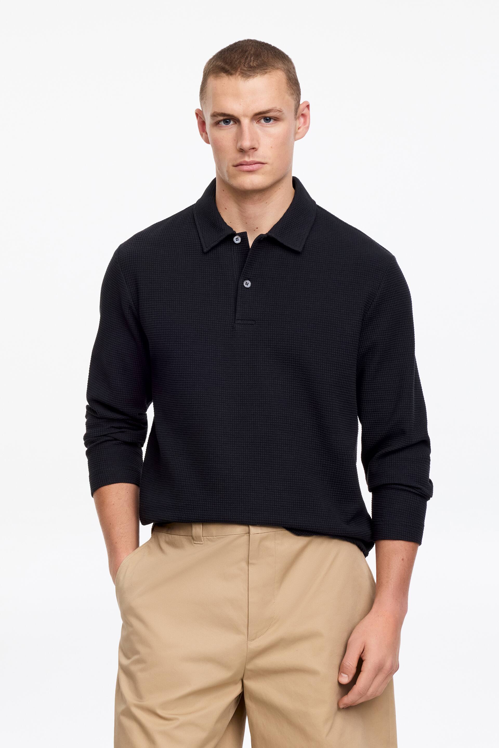 Agrandir l'image: Un polo noir à manches longues présente une texture en maille gaufrée, un col classique à pointes et une patte de boutonnage à deux boutons avec des boutons blancs, orientée vers l'avant.