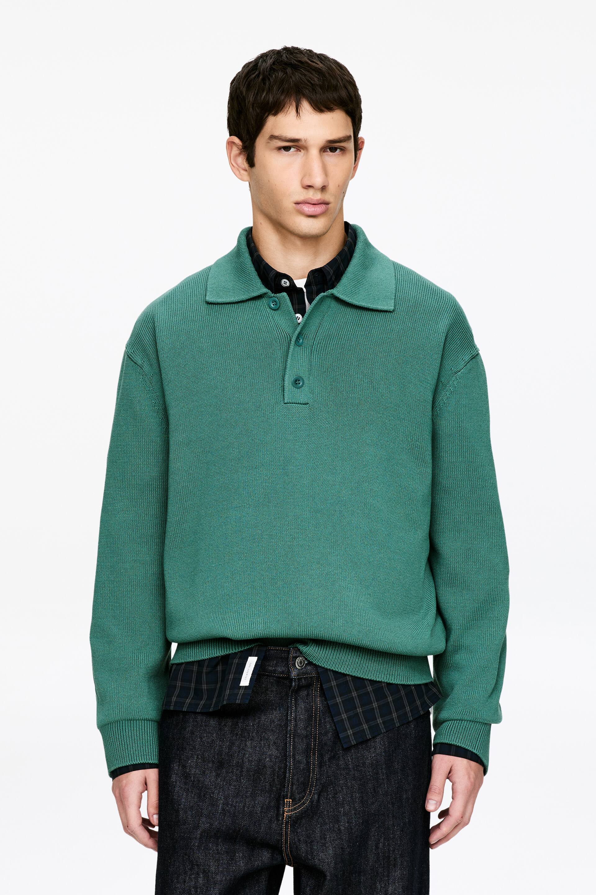 Ingrandisci l'immagine: Un maglione polo in maglia verde viene presentato frontalmente, caratterizzato da un colletto a coste aperto, un'abbottonatura a due bottoni, polsini a coste e un orlo a coste. È sovrapposto a una camicia a quadri scura con colletto.