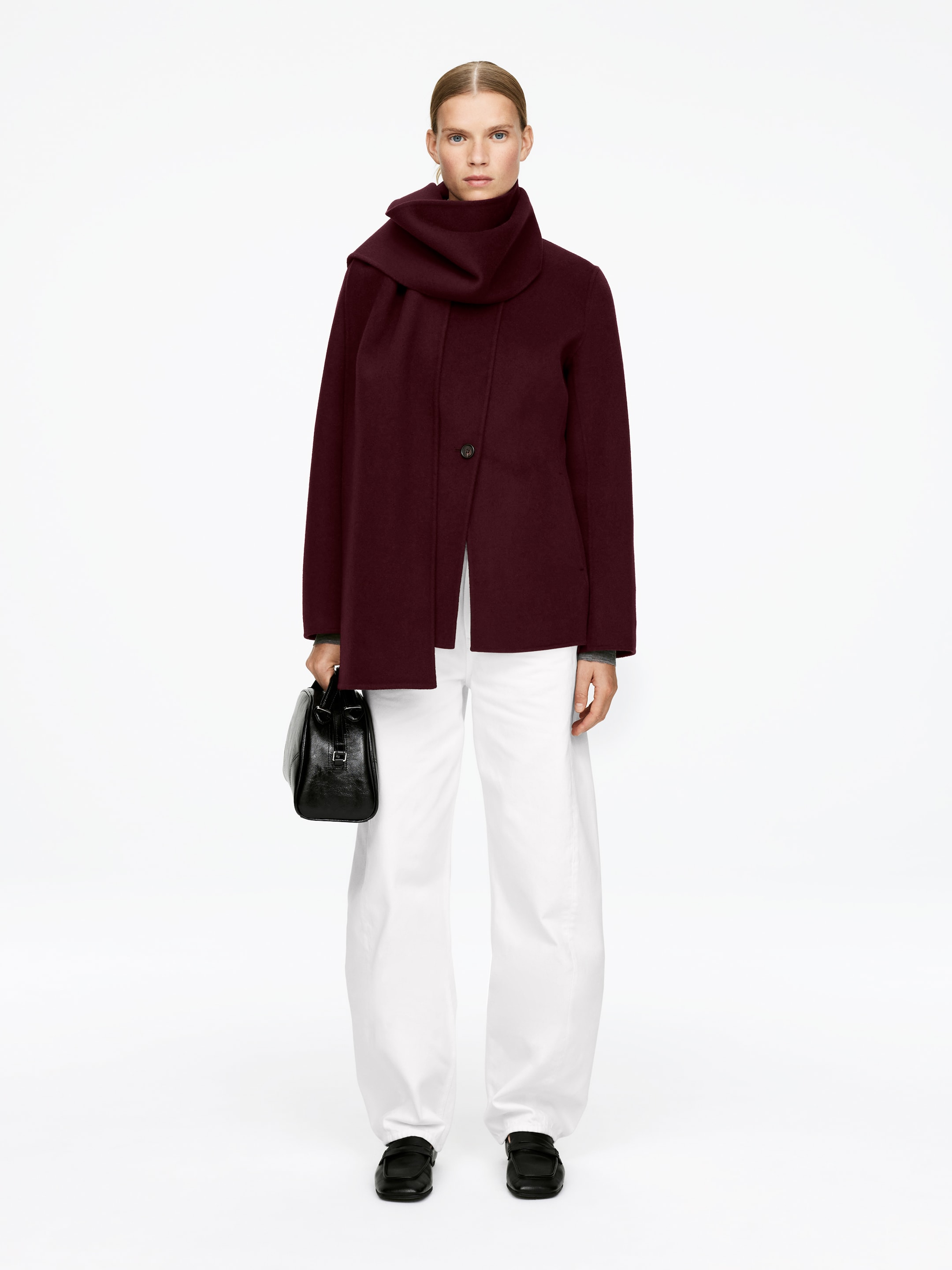 Agrandir l'image: Un manteau court bordeaux foncé présente une fermeture à bouton unique, décentrée, et un col montant, avec une écharpe volumineuse assortie drapée autour du cou. Le manteau est superposé à un pantalon blanc à jambes larges et un haut gris à manches longues. Des mocassins noirs sont portés, et un sac à main structuré noir est tenu.