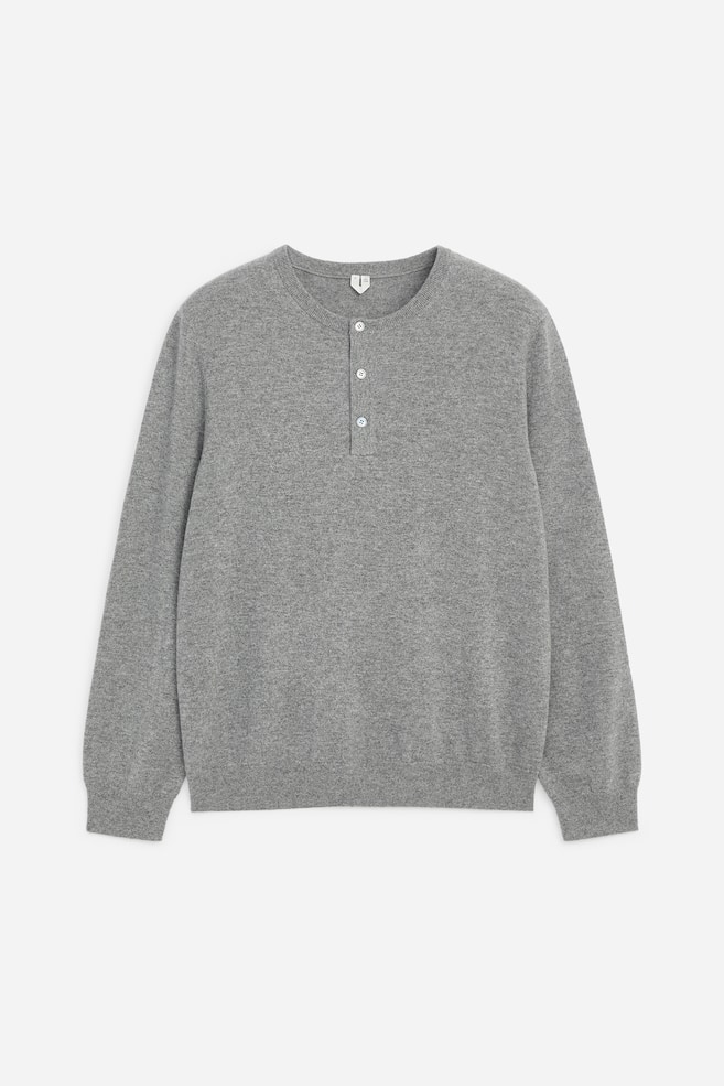Se muestra un jersey henley jaspeado gris claro desde el frente, con un escote de canalé de doble capa y una tapeta de tres botones con botones de color claro. La prenda de manga larga presenta una textura de punto fino y se remata con puños y bajo de canalé. - 1