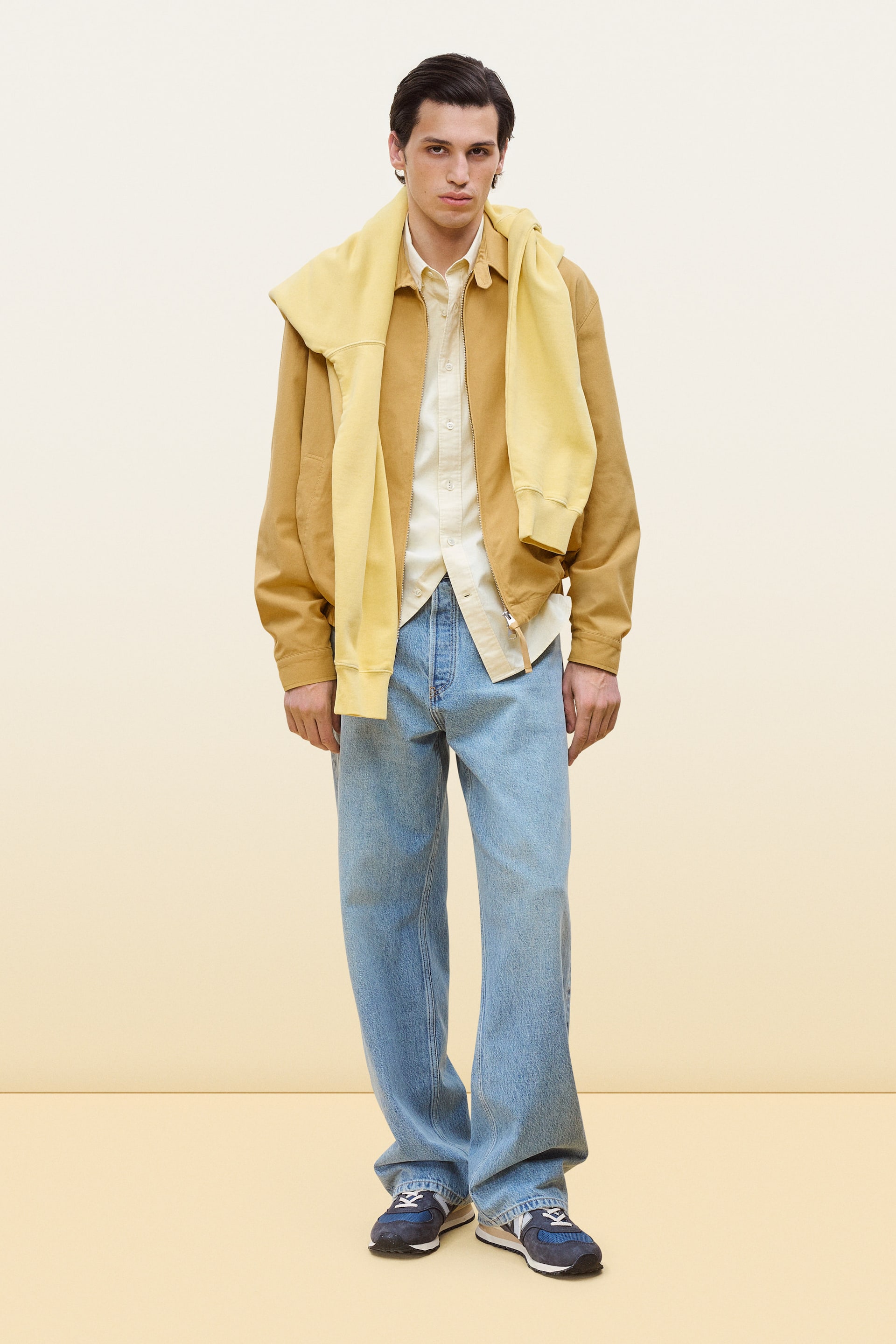 View larger image: A complete ensemble is showcased, featuring a tan utility jacket worn open over a cream button-up shirt. A light yellow crewneck sweatshirt is draped over the shoulders, complementing light wash wide-leg jeans and blue and beige trainers, all displayed from a front-facing angle.