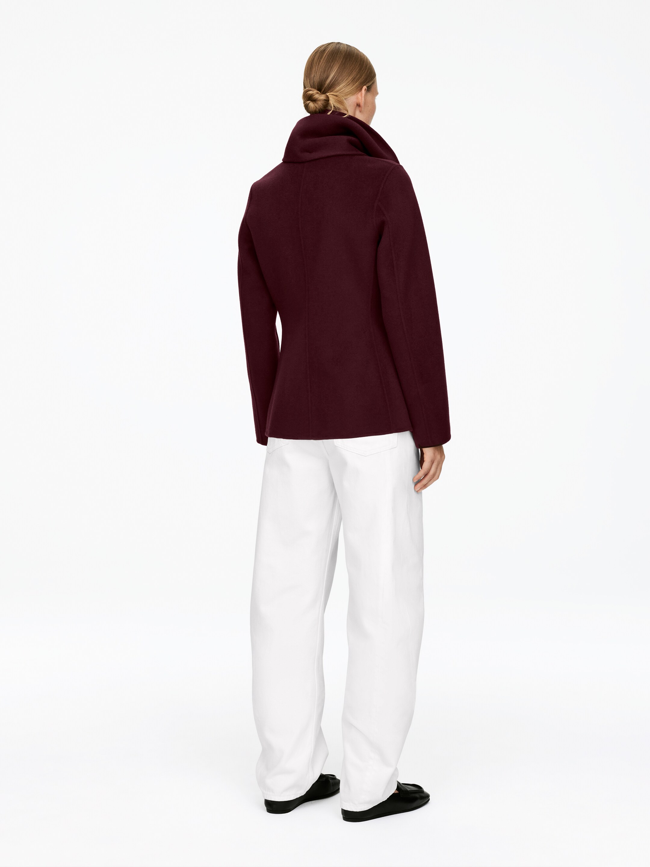 Agrandir l'image: Un manteau court bordeaux foncé est vu de dos, légèrement incliné vers la droite. Il présente un col montant, des manches longues et une coupe ajustée à travers le corps, semblant être fait d'un tissu structuré et se terminant à la hanche.