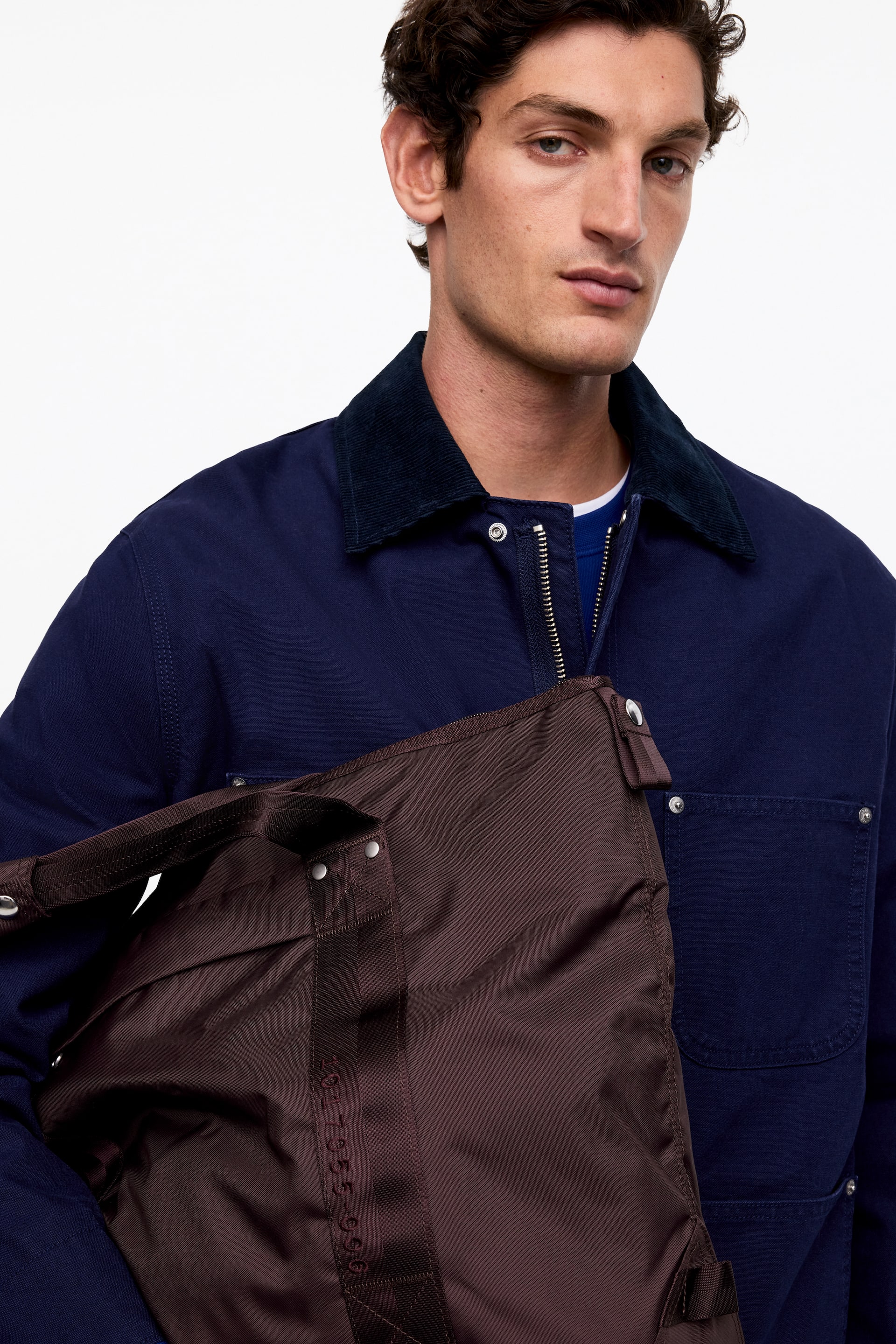 Größeres Bild ansehen: Eine dunkelbraune Nylon-Umhängetasche mit tonalen Stoffriemen und silberfarbenen Beschlägen wird vom Model getragen, der obere Reißverschluss ist teilweise geöffnet.