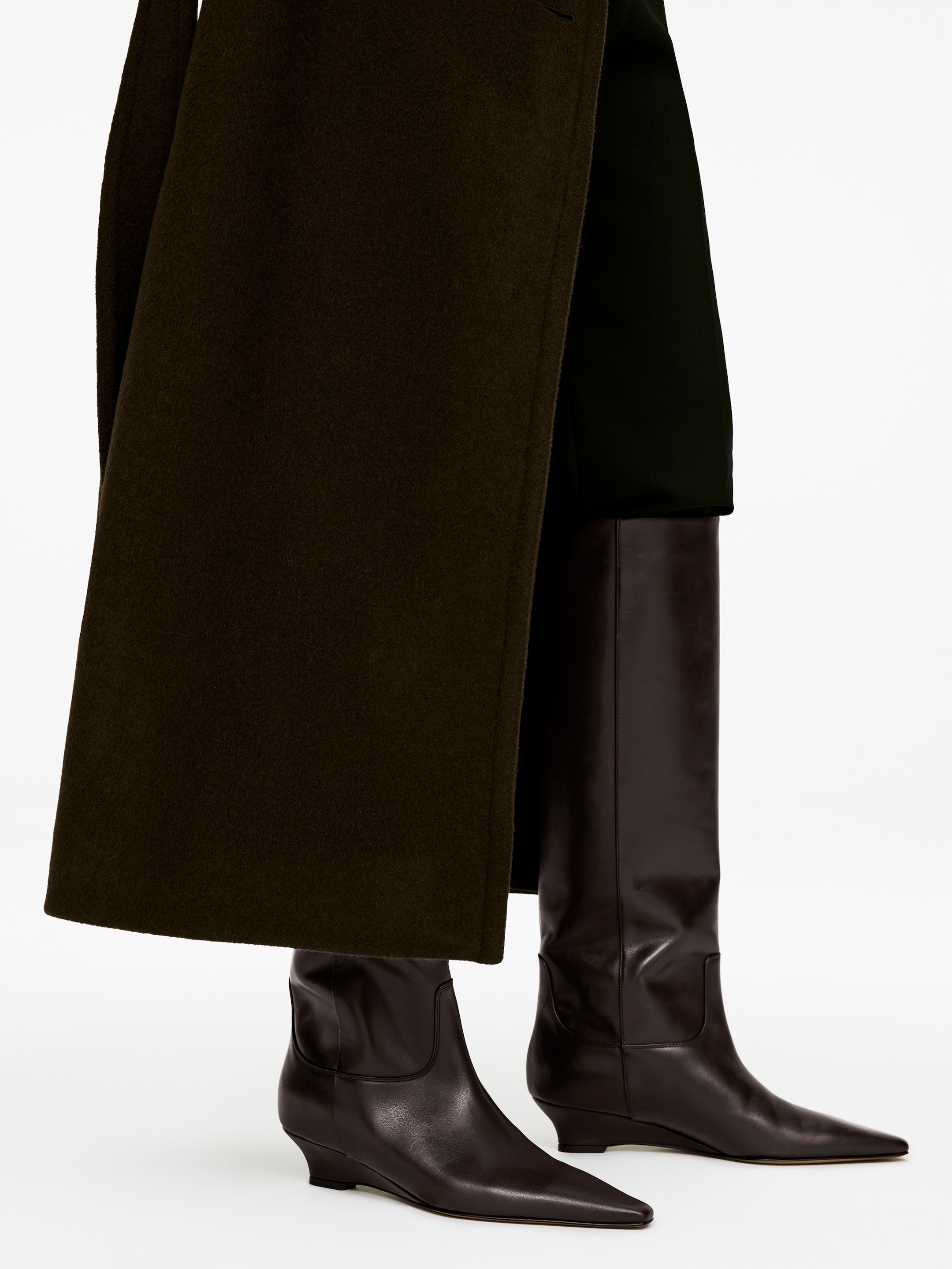 Agrandir l'image: Des bottes en cuir marron foncé montantes jusqu'aux genoux sont présentées légèrement de face, en perspective angulaire. Les bottes présentent des bouts pointus et un talon compensé bas, avec de longues tiges visibles. Un manteau olive foncé est partiellement drapé sur la partie supérieure des bottes.