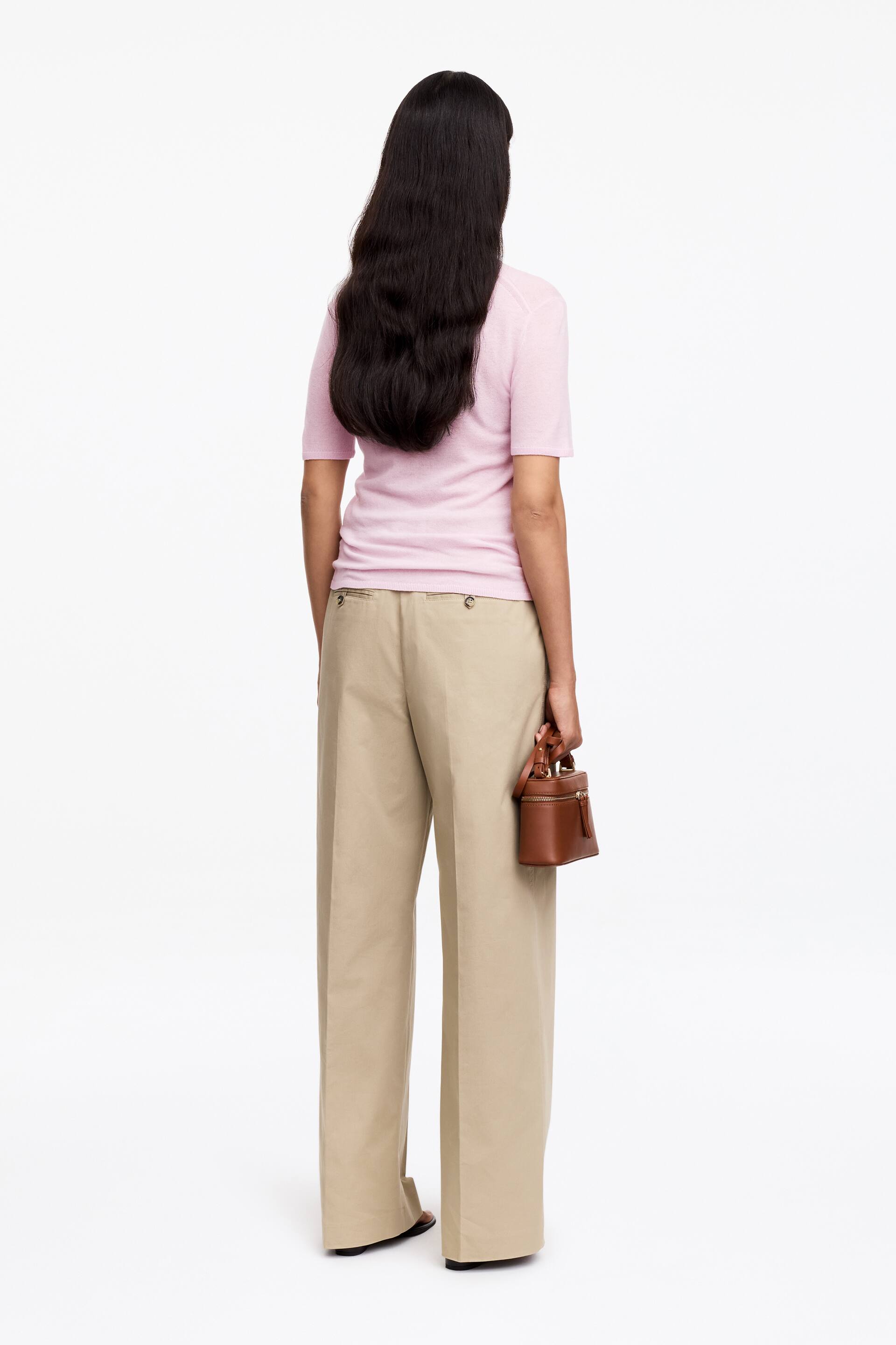 View larger image: A light pink short-sleeved knit top fits smoothly from the rear view, styled with high-waisted beige wide-leg trousers featuring a relaxed fit. A small brown structured handbag with a top zip is held by its handle.
