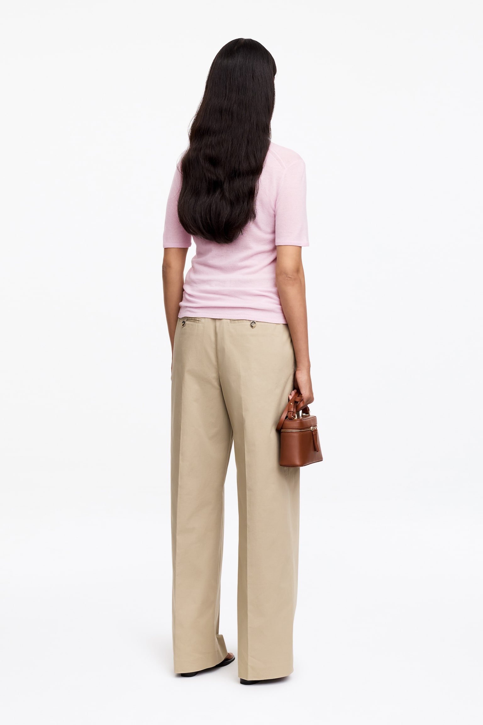Een lichtroze gebreide top met korte mouwen past soepel vanuit een achteraanzicht, gestyled met beige hoog getailleerde wijde broek met een ontspannen pasvorm. Een kleine bruine gestructureerde handtas met een bovenrits wordt vastgehouden aan het handvat. - 1