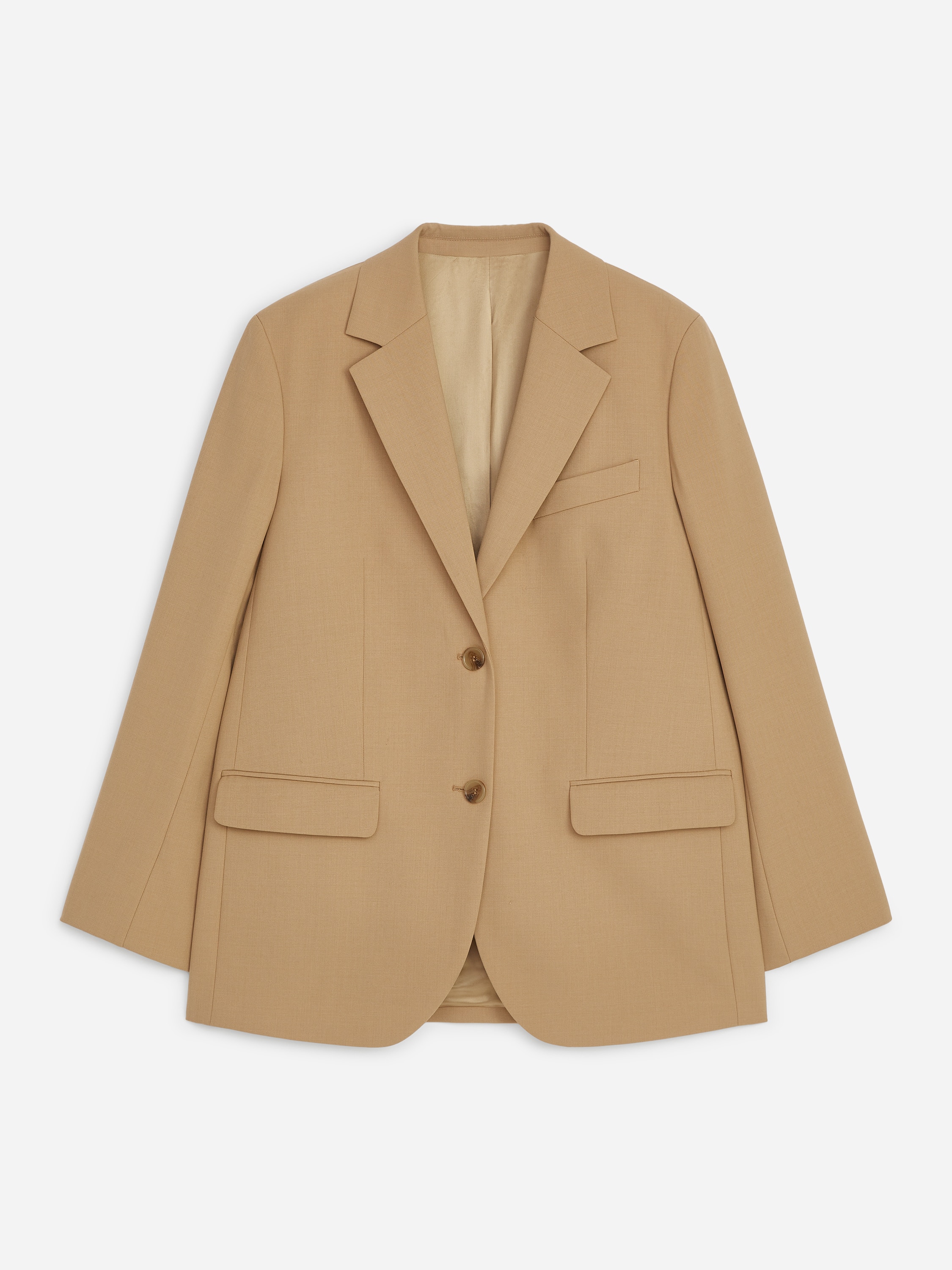 En beigefärgad, skräddarsydd blazer med avslappnad passform bärs av modellen och har slag med notching, en bröstficka med passpoal samt framfickor med lock. Blazern stylas med matchande byxor med vida ben, och modellen vilar ena handen i en byxficka.