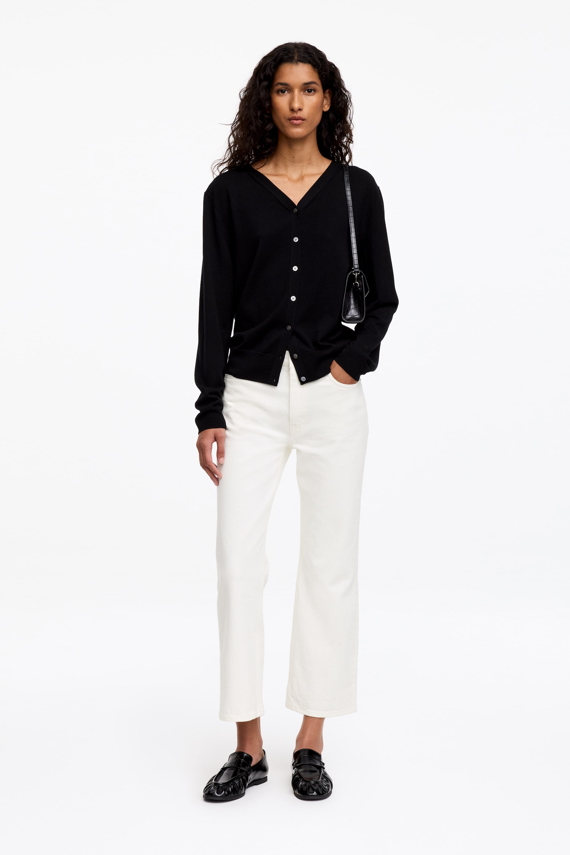 Vezi imaginea mai mare: Modelul poartă blugi albi cropped kick-flare și stă cu o mână băgată în buzunarul din față. Blugii slim-fit, cu talie medie, sunt asortați cu un cardigan negru cu nasturi, o geantă neagră de umăr și pantofi loafers negri.