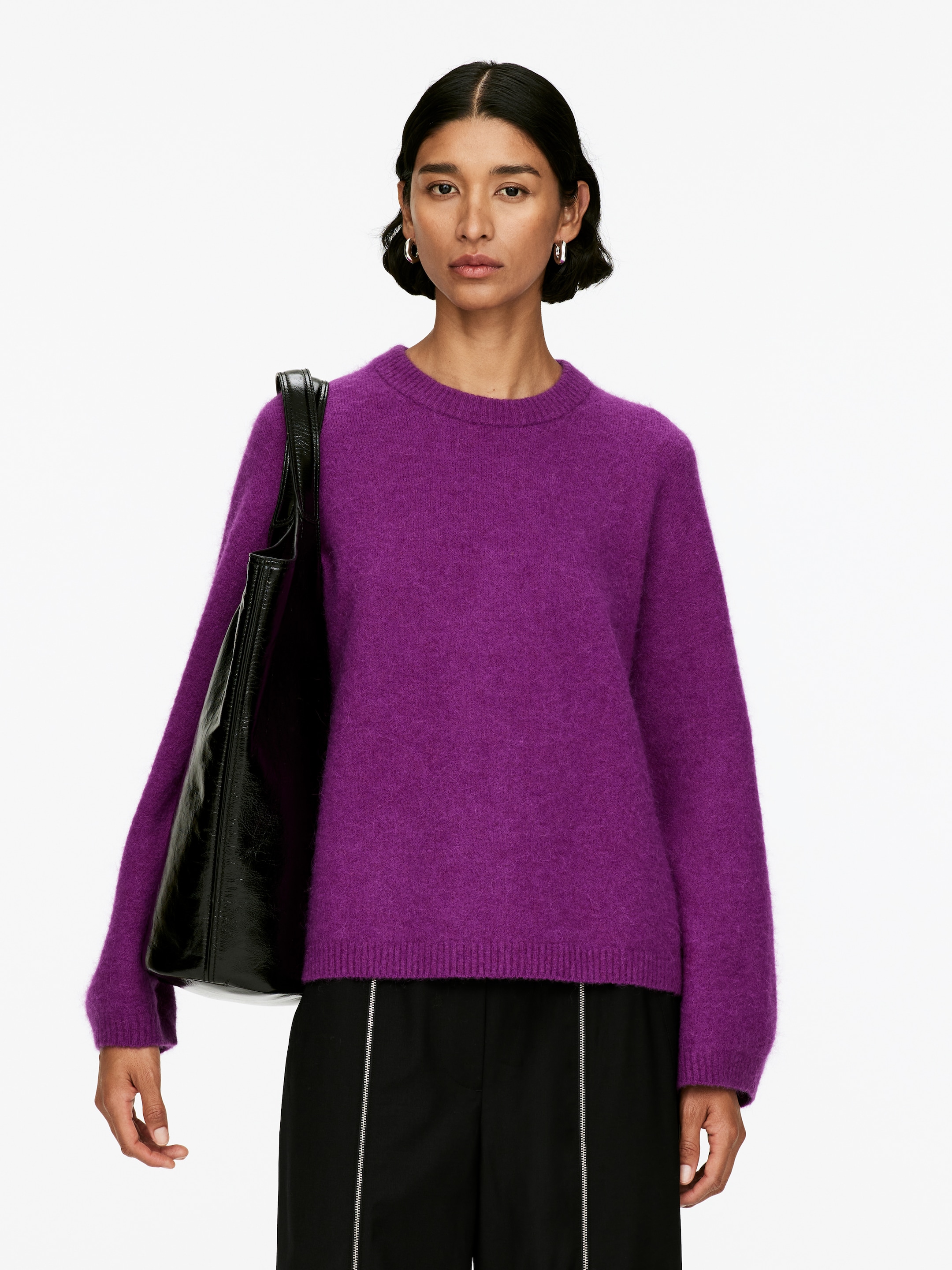 Agrandir l'image: Un pull à col rond violet foncé est présenté de face, avec une texture duveteuse et une coupe décontractée. Il présente des poignets et un ourlet côtelés, et un sac à bandoulière noir obscurcit partiellement sa manche gauche, tandis que des pantalons noirs sont visibles en dessous.
