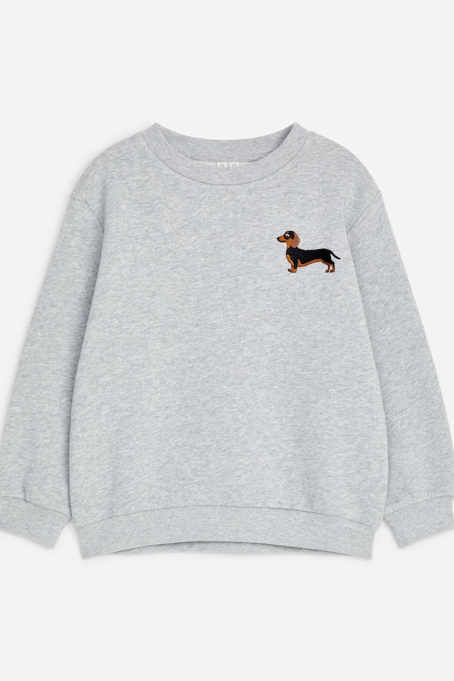 A light grey sweatshirt is presented in a relaxed fit with dropped shoulders, a ribbed crew neck, ribbed cuffs, and a ribbed hem from the front. A small embroidered black and tan dachshund details the upper left chest. - 1