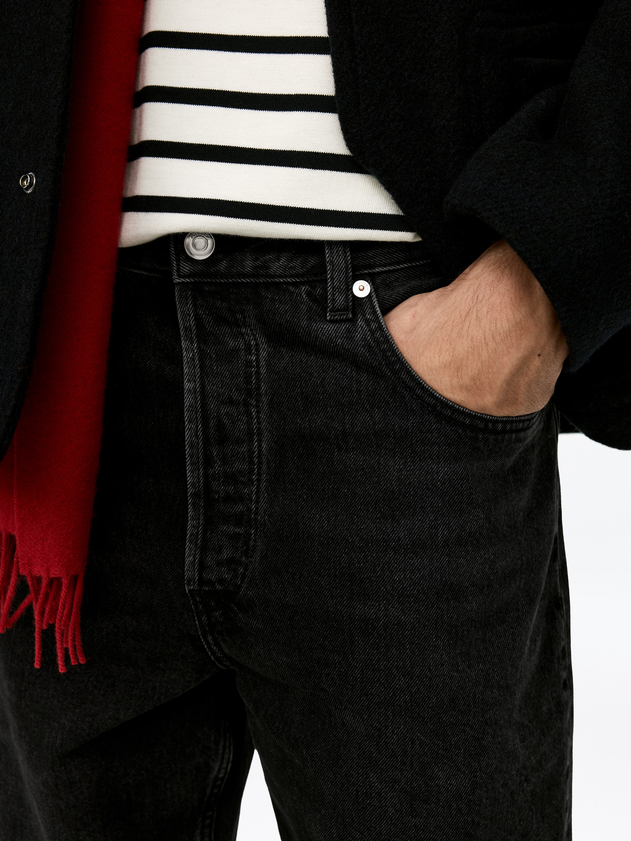 Grösseres Bild ansehen: Verblasste schwarze Waschdenimjeans werden aus einer oberen Vorderansicht präsentiert, mit einem Metallknopfverschluss und Nieten an den Vordertaschen. Eine Hand ruht in der rechten Vordertasche, während ein weiß-schwarz gestreifter Pullover in die Taille gesteckt wird, eine schwarze Jacke darüber getragen wird und ein roter Fransenschal an der Seite drapiert ist.