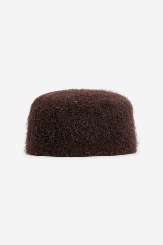 Ein dunkelbrauner, randloser Pillbox-Hut ist von vorne zu sehen und präsentiert eine stark strukturierte, flauschig gebürstete Wollaußenseite mit einer geformten, kuppelförmigen Silhouette. - 1