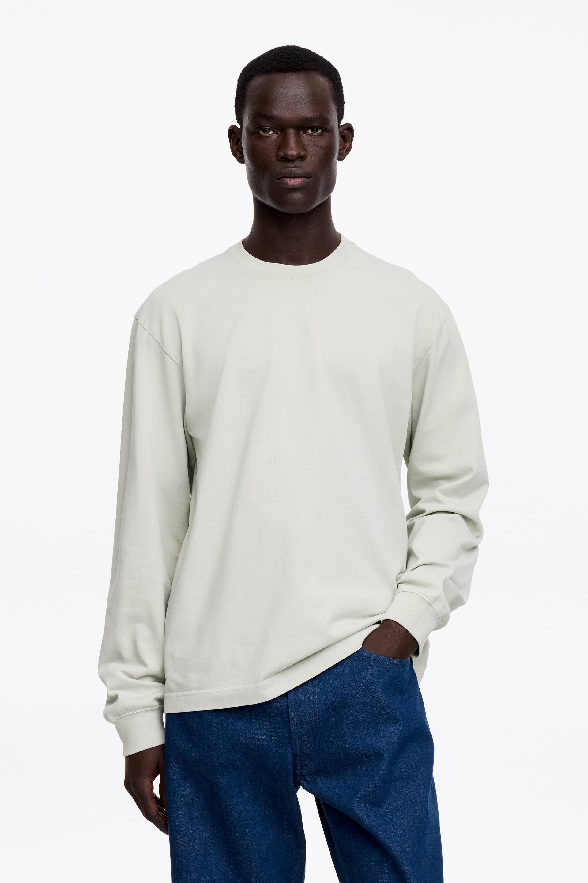 Ver imagen más grande: Una camiseta de manga larga de color verde claro, confeccionada en algodón y con un clásico cuello redondo y mangas con puños, está siendo llevada por el modelo. El modelo está de pie con una mano descansando en el bolsillo de unos vaqueros azul oscuro.