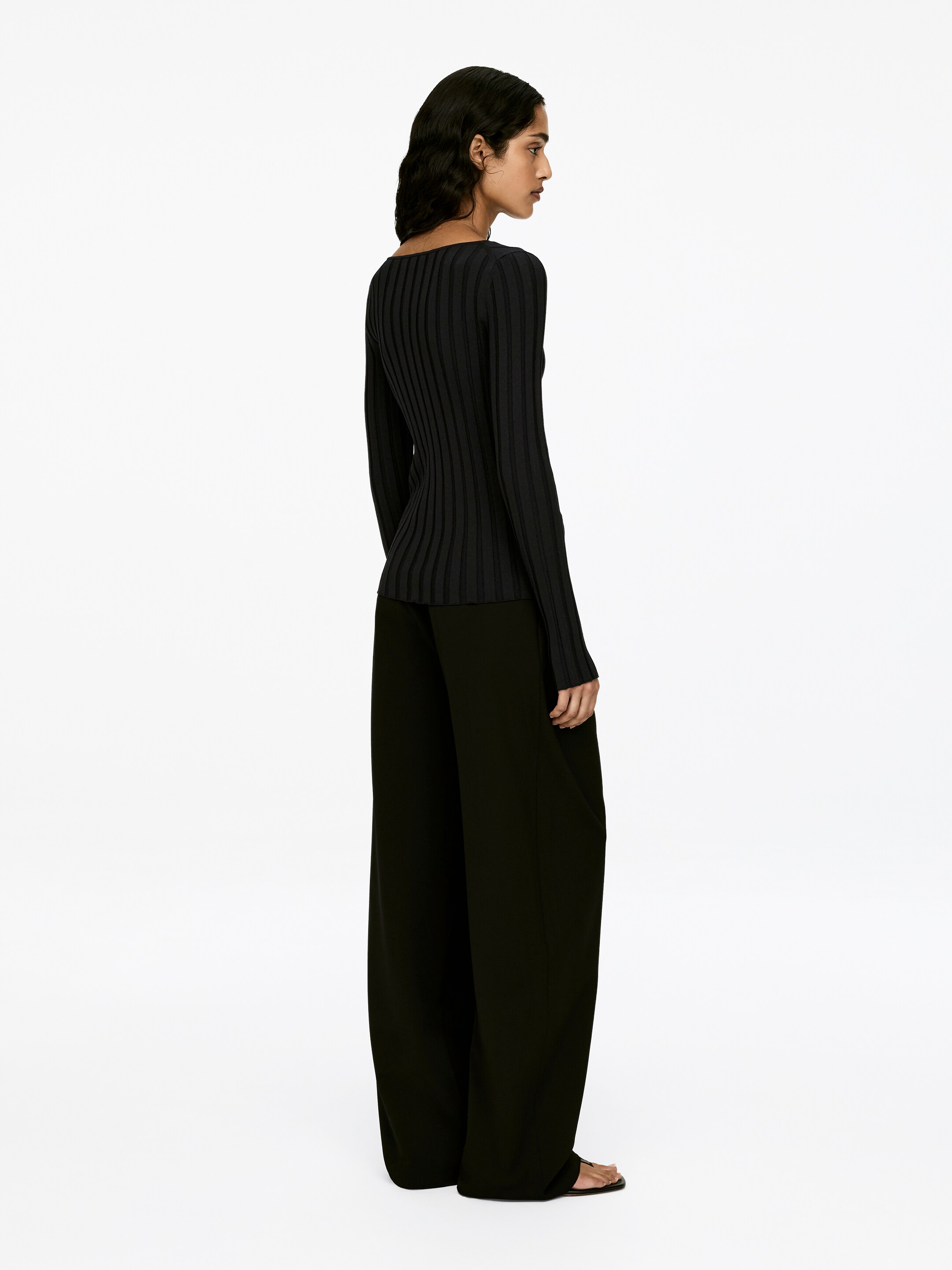 View larger image: A dark, slim-fit, long-sleeved jumper is exhibiting a prominent rib-knit texture as it is being worn by the model. The garment is being styled with dark, wide-leg trousers.