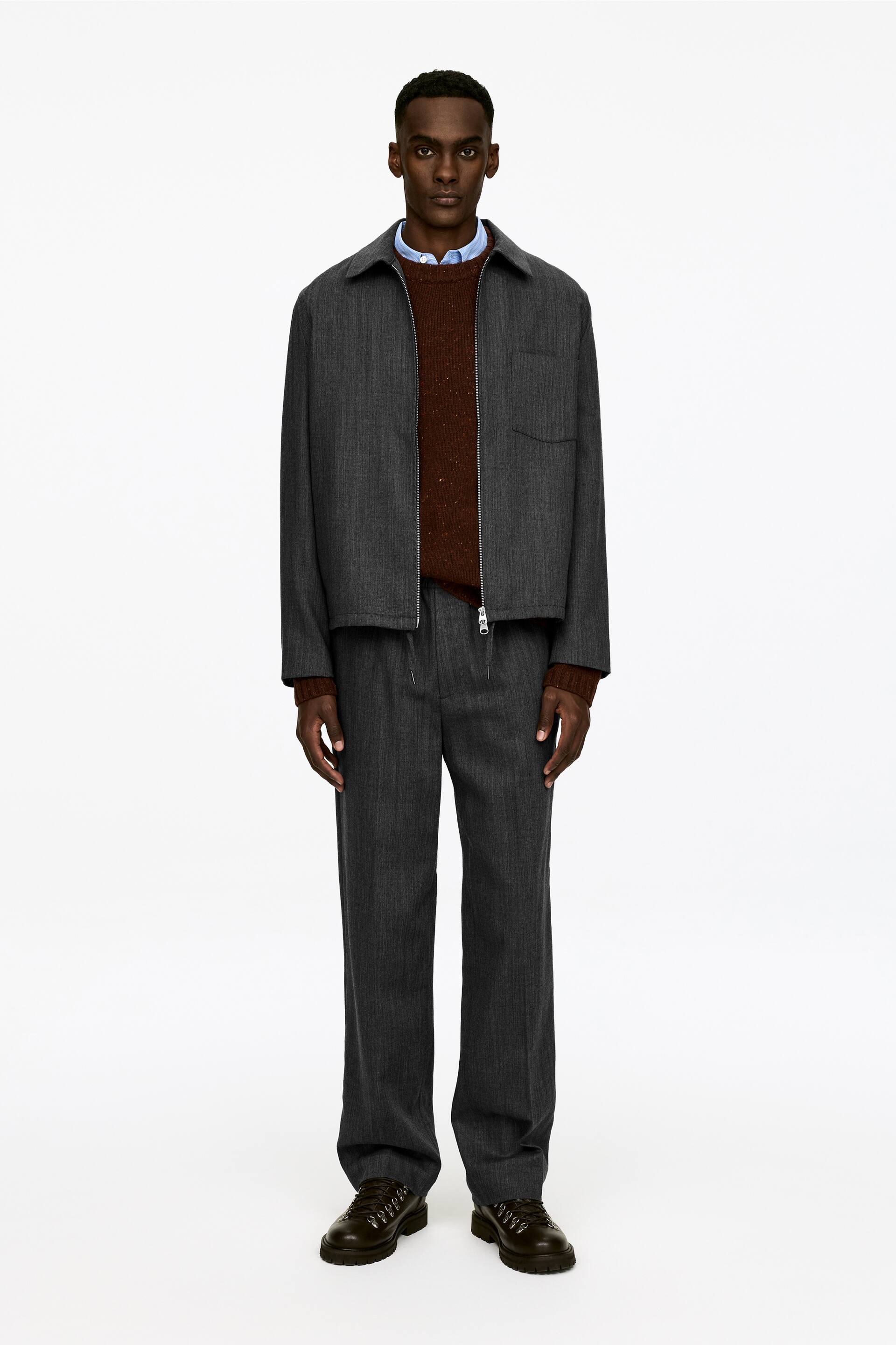 Преглед на по-голямо изображение: Сив комплект от две части, включващ яке с яка и цип отпред с джоб на гърдите и панталони с прави крачоли, е представен отпред. Якето е слоено върху кафяв плетен пуловер и светлосиня риза с яка, а тъмнокафяви обувки с връзки завършват аутфита.