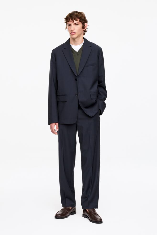 A dark navy suit is being showcased, featuring a two-button single-breasted jacket with notch lapels and flap pockets, paired with matching wide-leg trousers. A green V-neck jumper over a white t-shirt is layered underneath the jacket, and brown leather loafers are being worn with the trousers. - 1