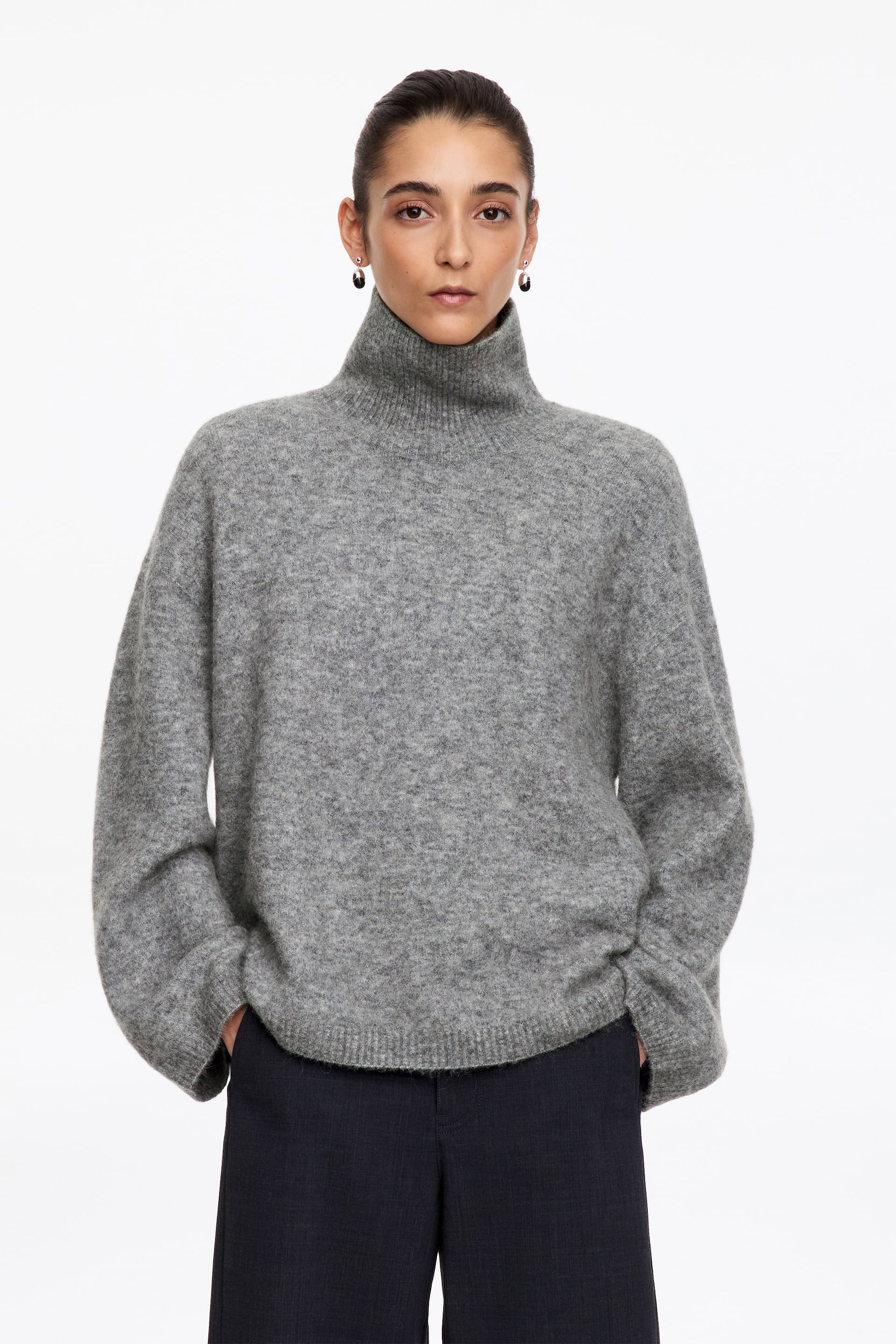 Ver imagen más grande: La modelo lleva un jersey de cuello cisne gris, de textura afelpada, con silueta oversize y acabados de canalé. La prenda de punto se combina con unos pantalones oscuros de pernera ancha.