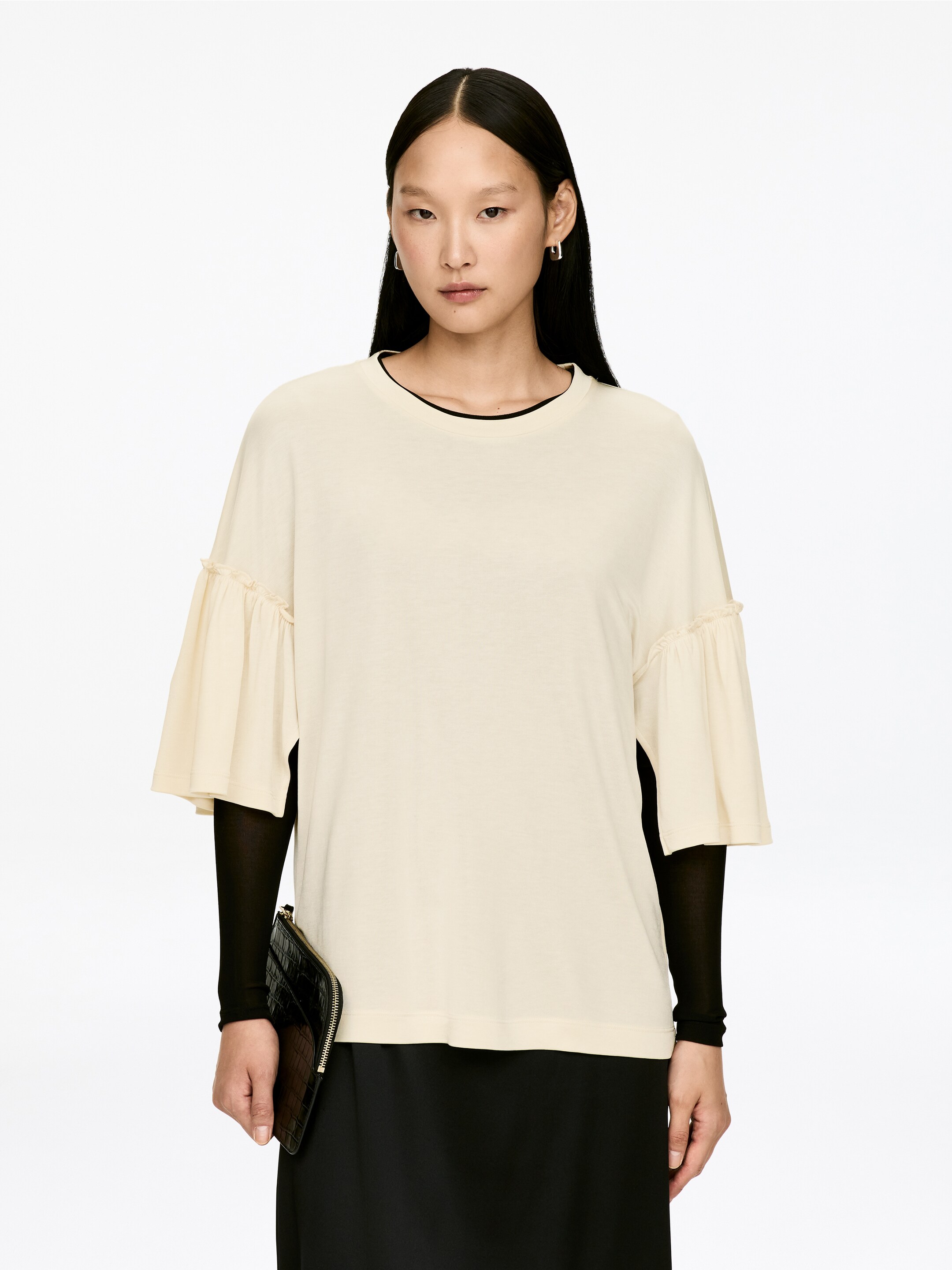 Visa större bild: En arbetsmodell med en krämfärgad, normal passform, rundhalsad T-shirt med volangärmar bärs av modellen över en svart långärmad topp. Modellen håller i en svart clutchväska.