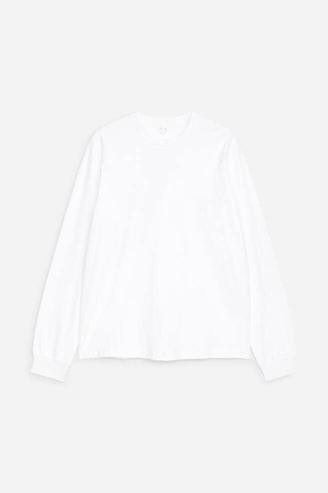 A white long-sleeve cotton T-shirt is displayed front-facing and laid flat, showcasing a loose fit, dropped shoulders, a ribbed crew neck, and ribbed cuffs. - 1