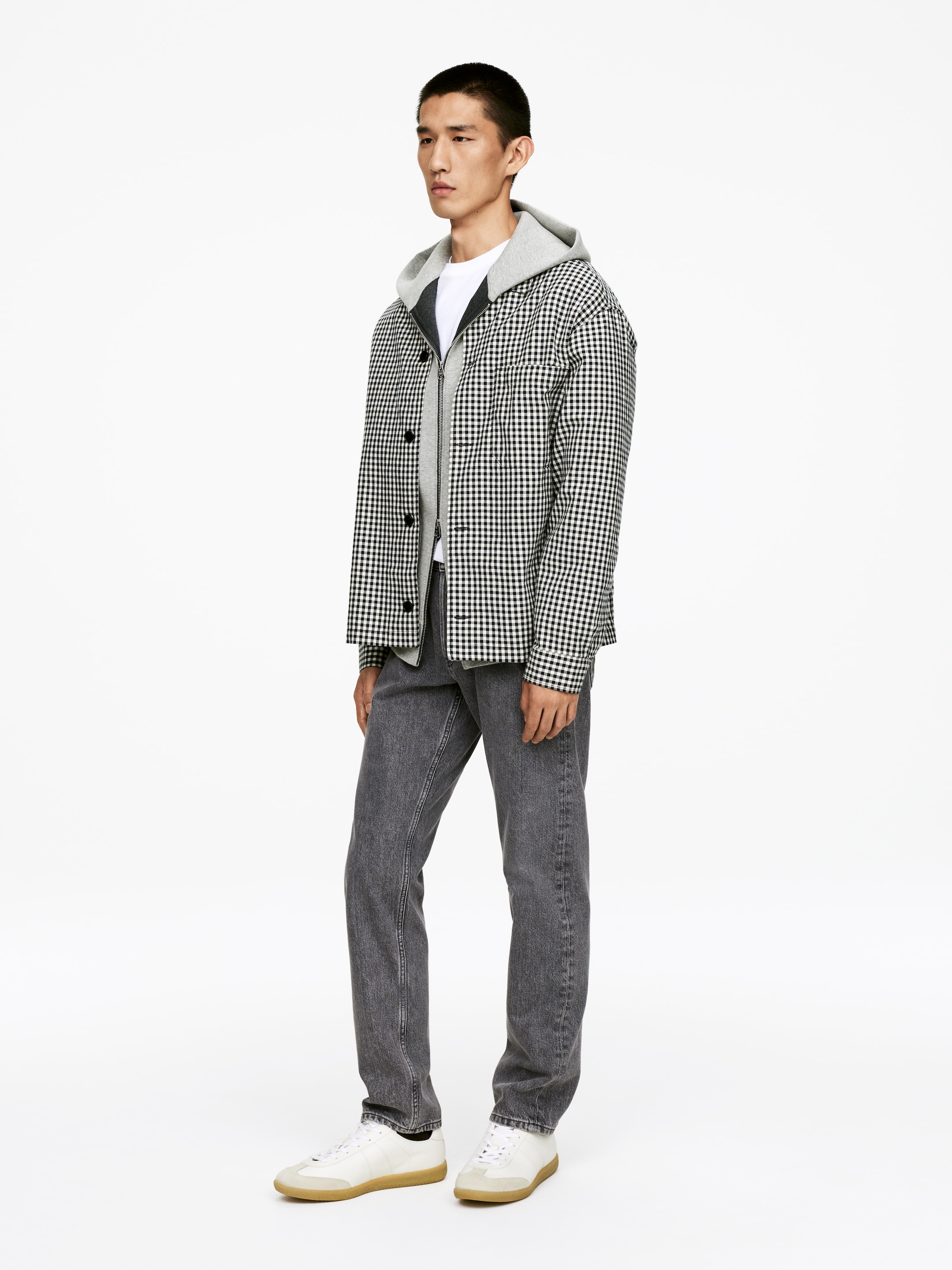 Grösseres Bild ansehen: Eine schwarz-weiß karierte Hemdjacke wird offen über einem grauen Kapuzenpullover getragen, der über einem weißen T-Shirt geschichtet ist. Dunkelgraue gerade geschnittene Jeans werden mit weißen Low-Top-Sneakers mit hellbraunen Gummisohlen getragen.