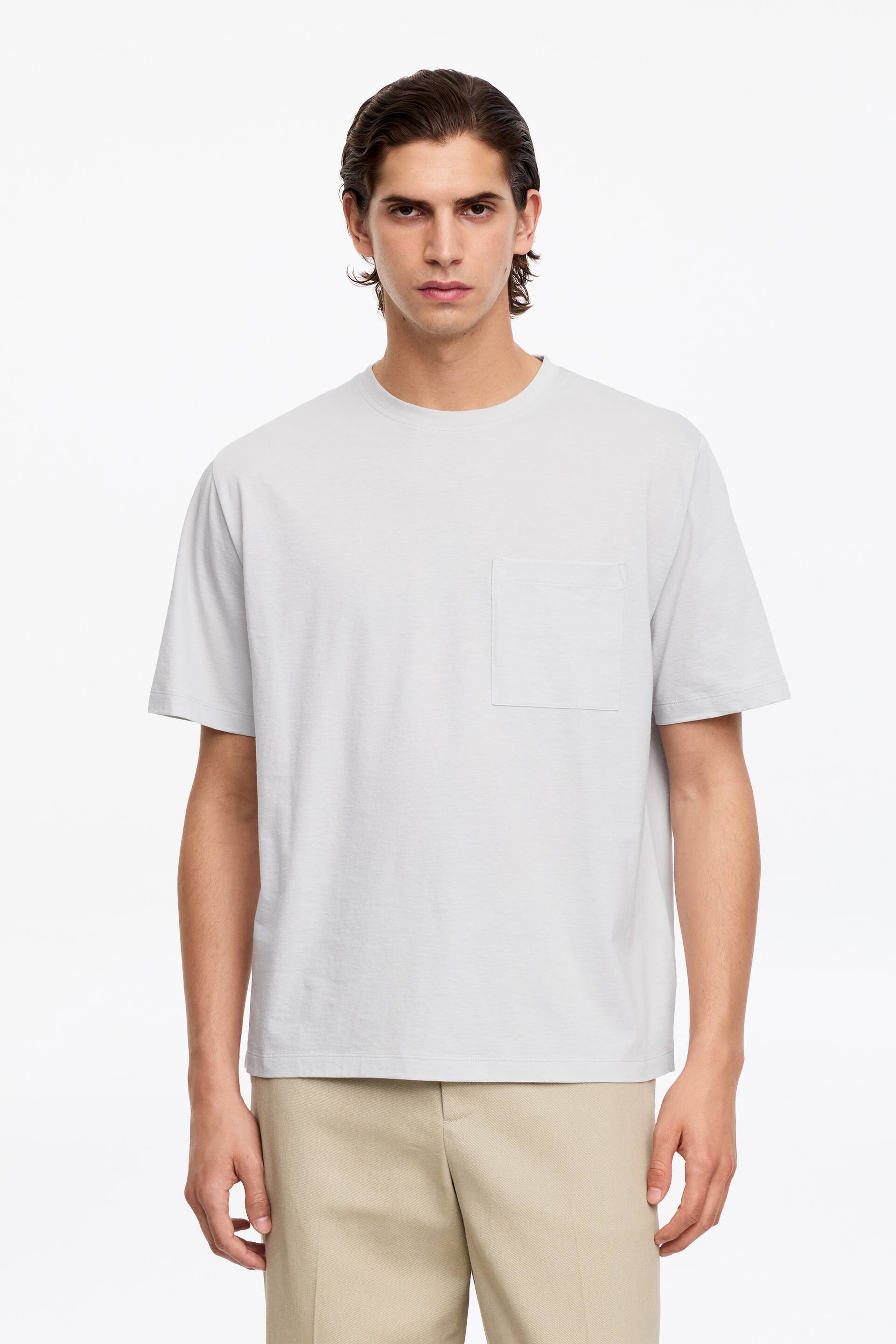 Zobacz większy obraz: Jasnoszara koszulka z krótkim rękawem skierowana przodem, z okrągłym dekoltem i pojedynczą kieszonką na lewej piersi. Prezentuje luźny krój.