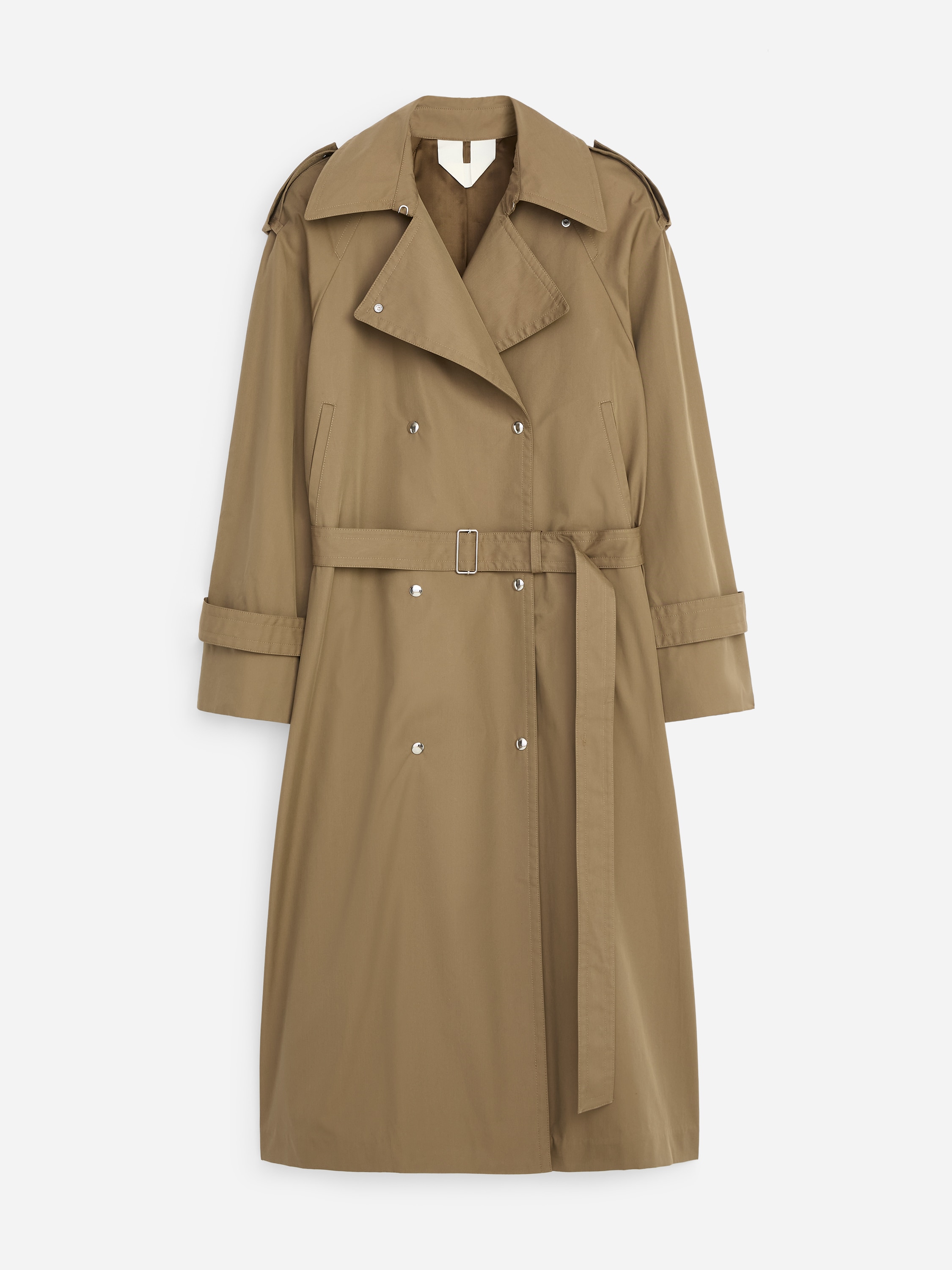 Ein olivgrüner doppelreihiger Trenchcoat wird von dem Modell getragen, das mit den Händen in den Seitentaschen steht. Der Mantel ist mit breiten Revers, Epauletten, Sturmklappen und silbernen Knöpfen gestaltet und wird über einem weißen Hemd, schwarzen Hosen und schwarzen Sandalen gestylt.