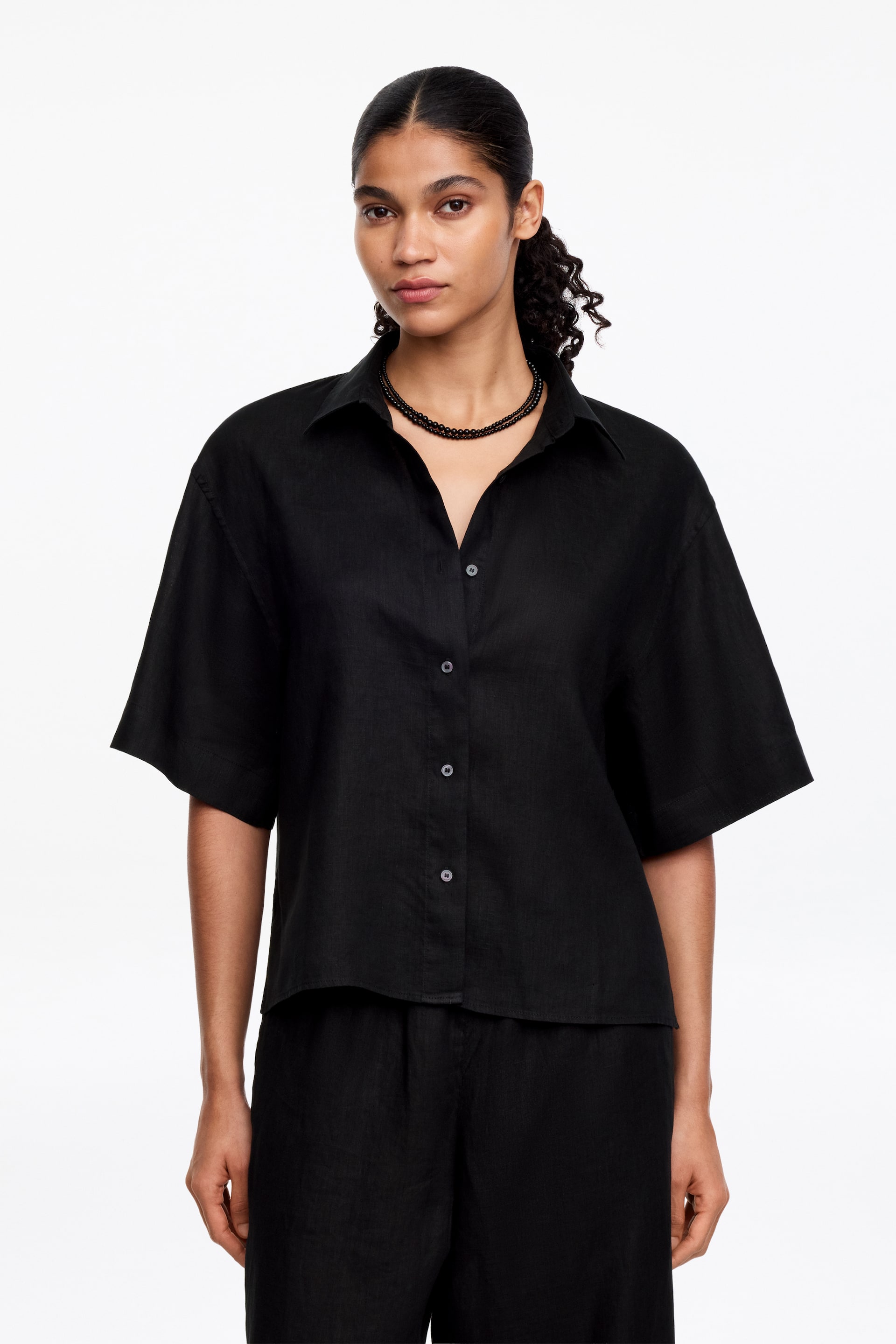 Agrandir l'image: Une chemise noire à manches courtes est présentée de face, avec un col pointu, des boutons sombres sur le devant et une coupe décontractée dans un tissu texturé. Elle est stylisée avec un pantalon noir assorti et un collier de perles noires.