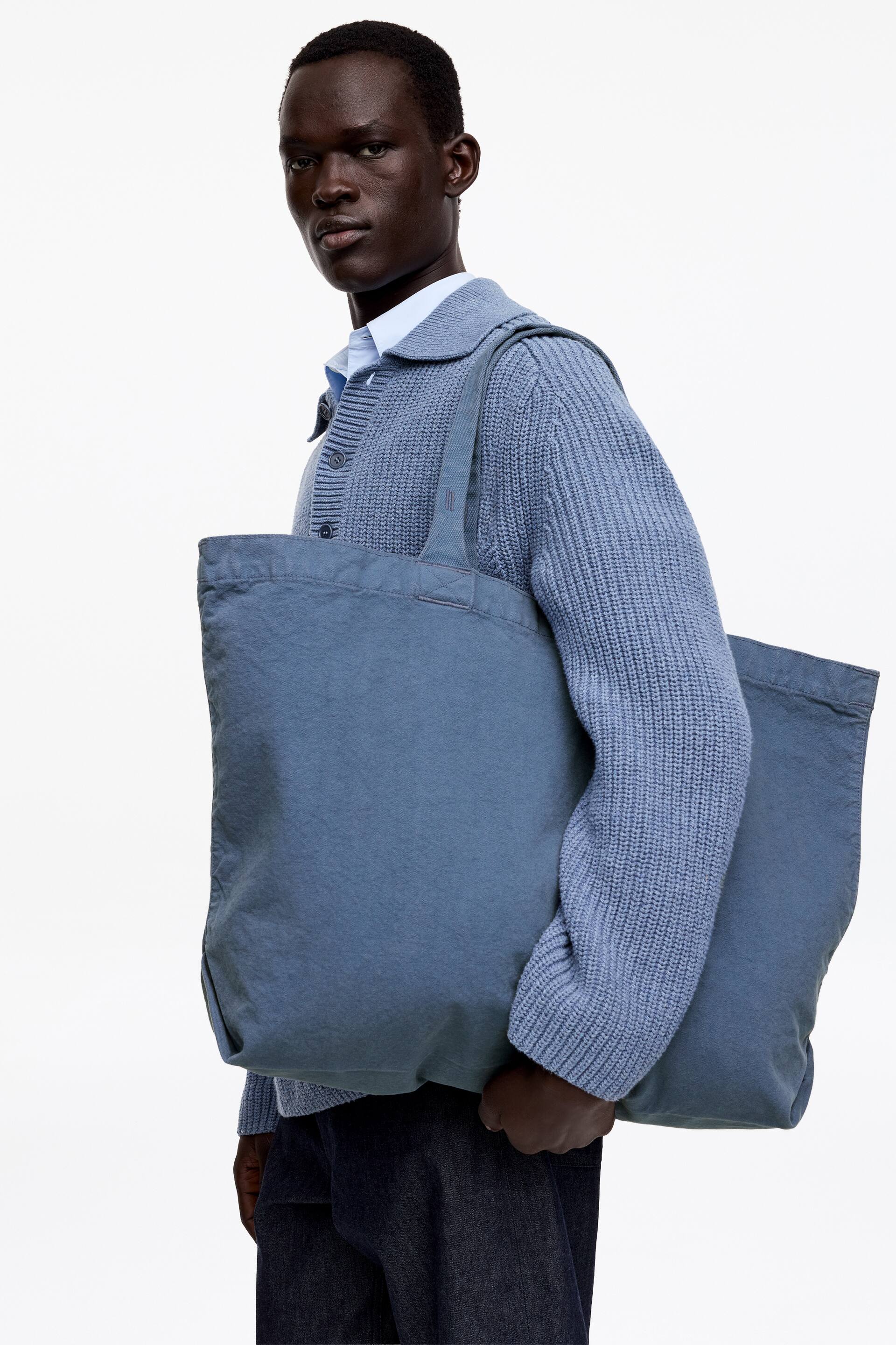 Näytä suurempi kuva: Liuskekiven sininen olkalaukku kannetaan olalla, esitellen sen suorakulmaista muotoa ja pehmeää, rypistettyä kangasta. Laukun kaksi leveää kahvaa ulottuvat sen yläreunasta, ja niissä on näkyvät tikkaukset.