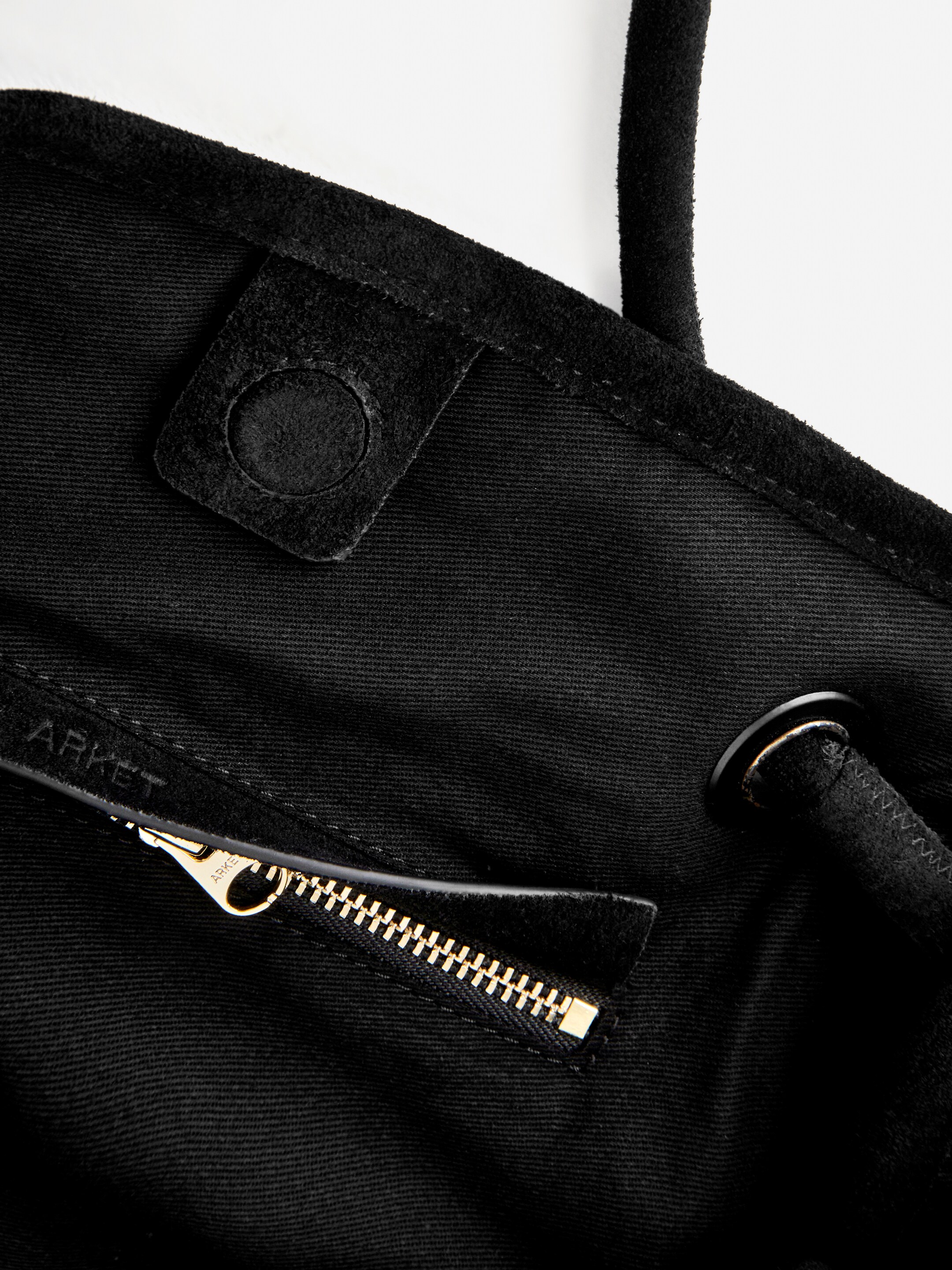 Agrandir l'image: Une vue rapprochée de l'intérieur d'un sac fourre-tout en daim noir montre une poche ouverte avec une fermeture éclair dorée, dont le tireur est embossé avec 'ARKET'. Une sangle tubulaire noire passe à travers un œillet noir avec un bord doré, à côté d'un rabat en daim noir avec un détail circulaire.