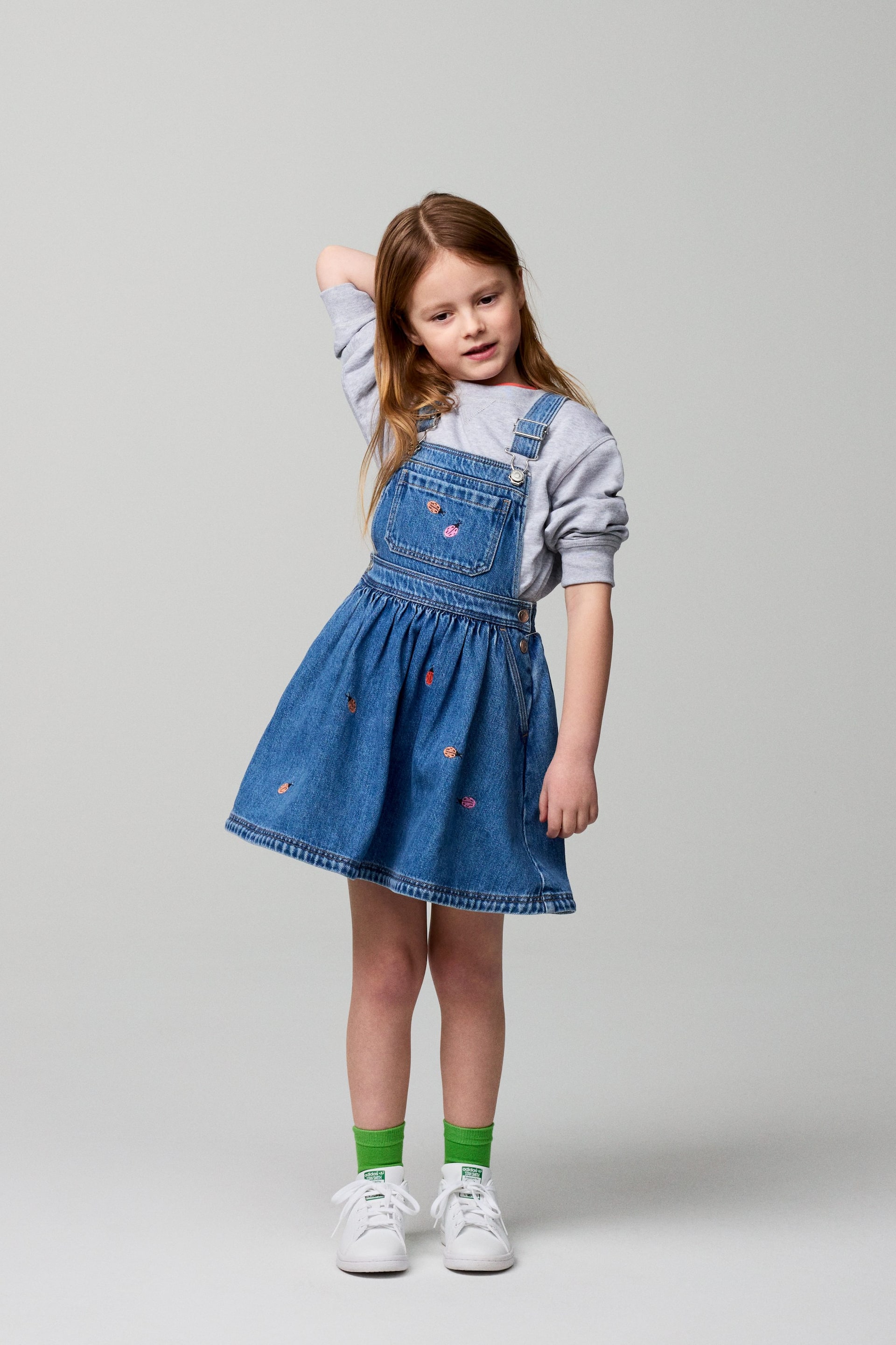 Προβολή μεγαλύτερης εικόνας: Ένα μπλε τζιν φόρεμα σαλοπέτα παρουσιάζεται, φορεμένο πάνω από ένα γκρι μακρυμάνικο τοπ. Διαθέτει μπροστινό τμήμα με τσέπη, ρυθμιζόμενες τιράντες ώμου με μεταλλικές αγκράφες και μια φούστα με σούρες που είναι διακοσμημένη με πολύχρωμα κεντημένα μοτίβα.