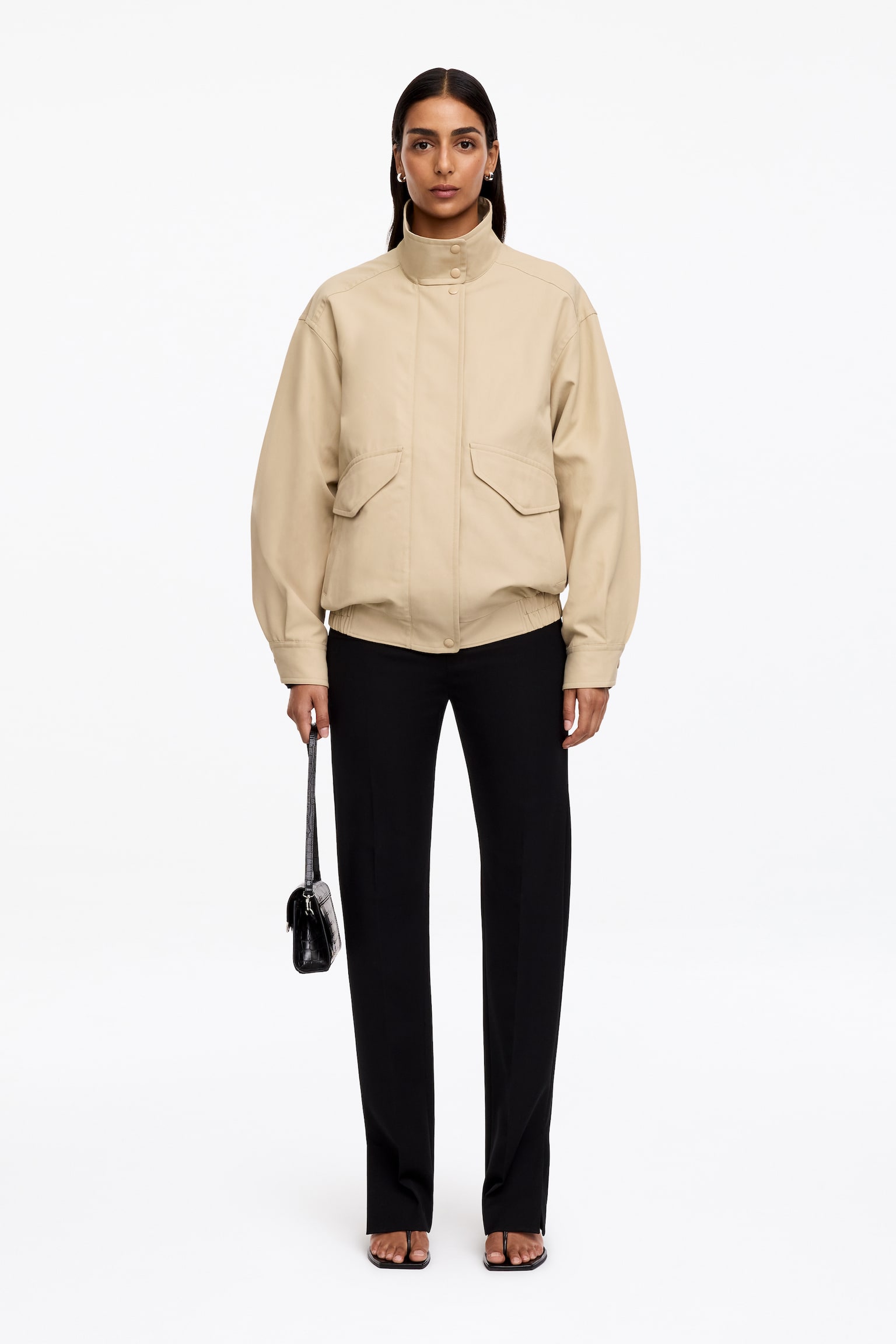 Béžová bunda Harrington je zobrazena ZEPŘEDU (FRONT) a má stojací límec se zapínáním na patentky, výrazné prošití na ramenou a přední légu skrývající zip, z něhož je u spodního okraje viditelný jezdec. Je navržena s dvěma velkými našitými kapsami s klopami zapínanými na knoflíky, manžetami zapínanými na knoflíky a elastickým vsazením v bočních spodních lemech. - 1