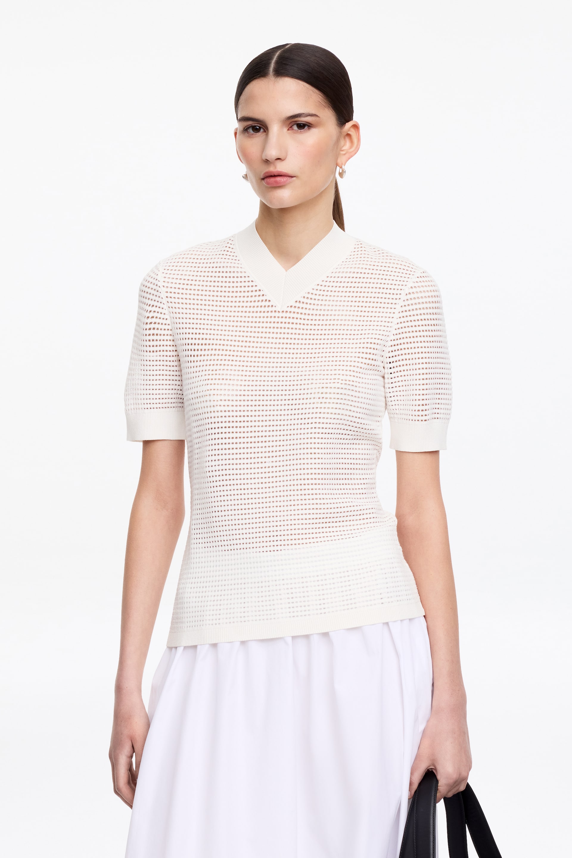Visa större bild: En vit öppenstickad topp har en V-ringning med ribbad krage och korta ärmar med ribbade muddar. Den har ett enhetligt perforerat mönster över hela huvudpartiet och en ribbad fåll.