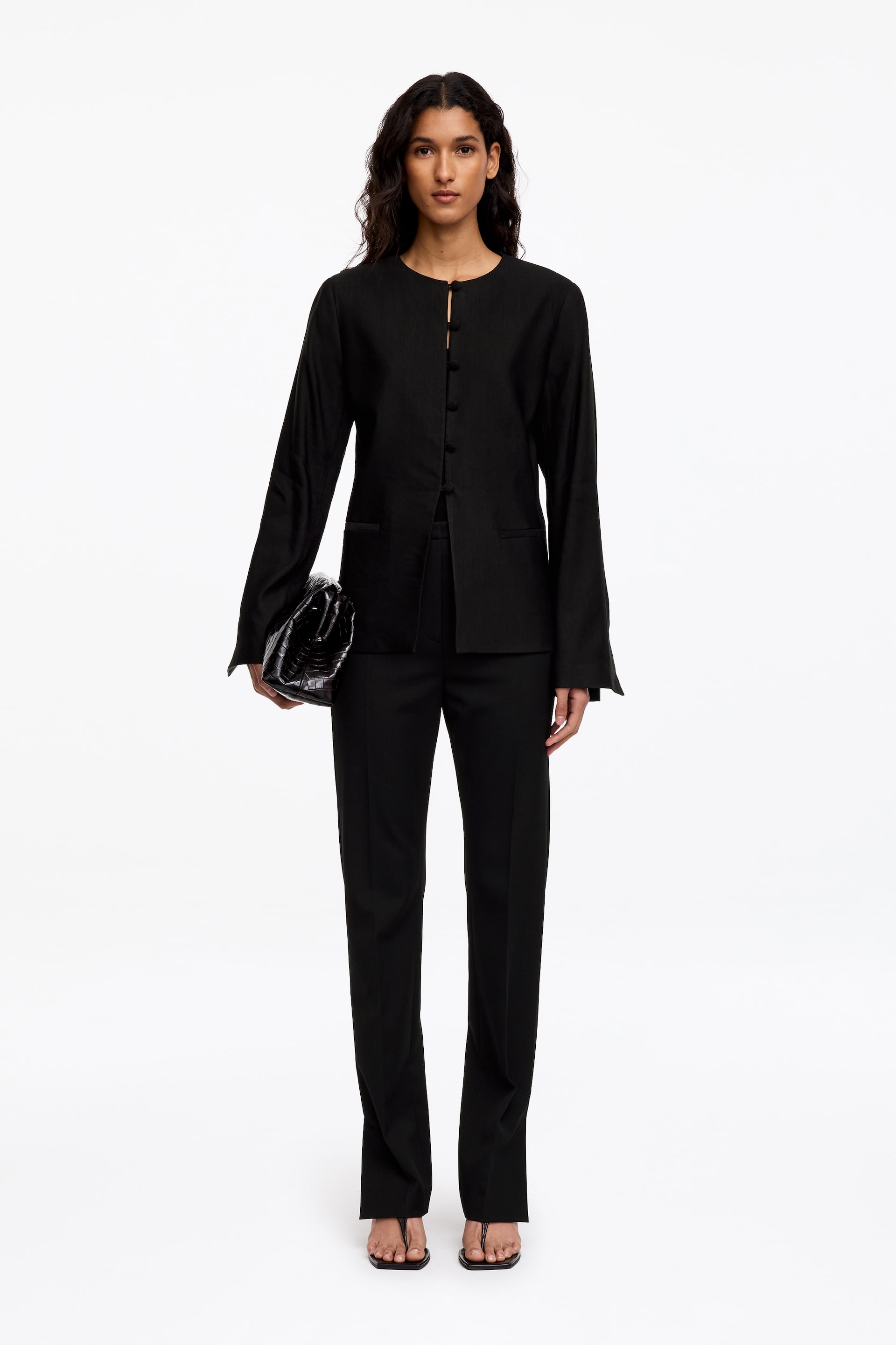 Größeres Bild anzeigen: Das Model trägt eine schwarze kragenlose Jacke mit Rundhalsausschnitt und vier Knöpfen vorne sowie eine schwarze Hose, schwarze minimalistische Sandalen und eine schwarze Clutch mit strukturierter Oberfläche.