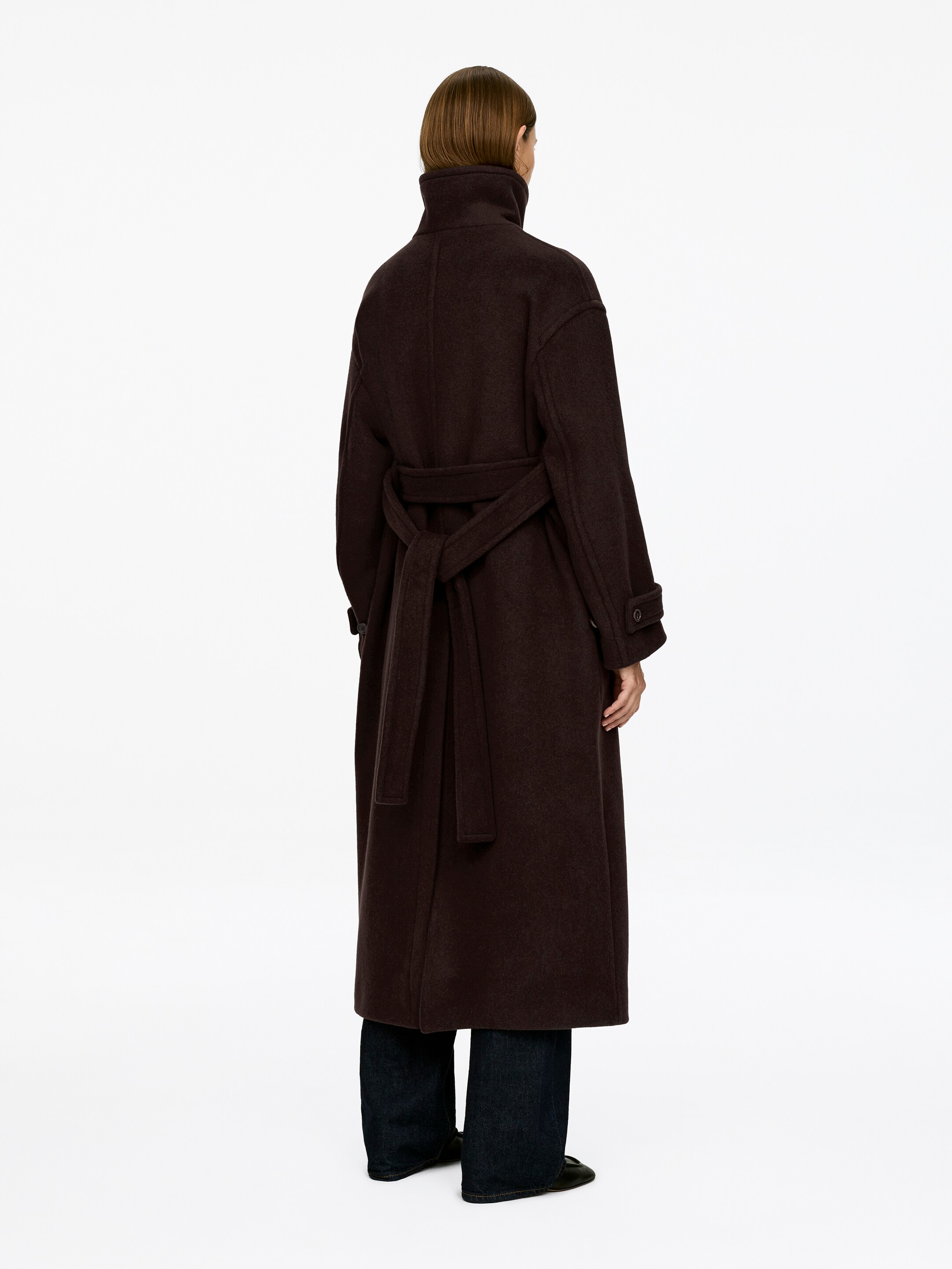Ver imagem maior: Um casaco maxi castanho escuro oversized com cinto está a ser usado pela modelo, visto de costas. O casaco tem uma gola alta e abas de manga com fechos de botão, e o seu cinto está atado nas costas.